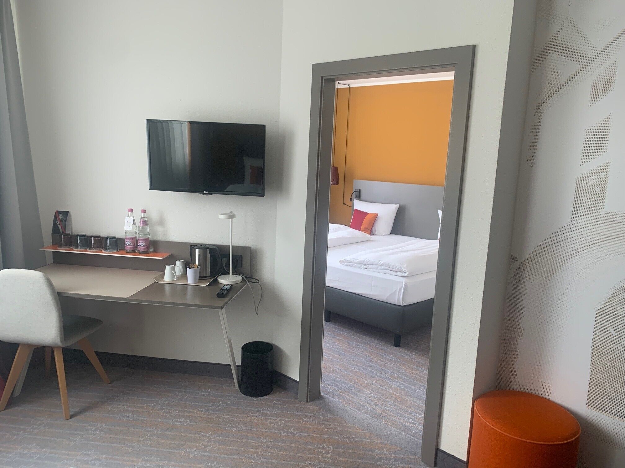 Quarto de hotel com cama, escrivaninha com televisão, cadeira e um pequeno cesto de lixo.