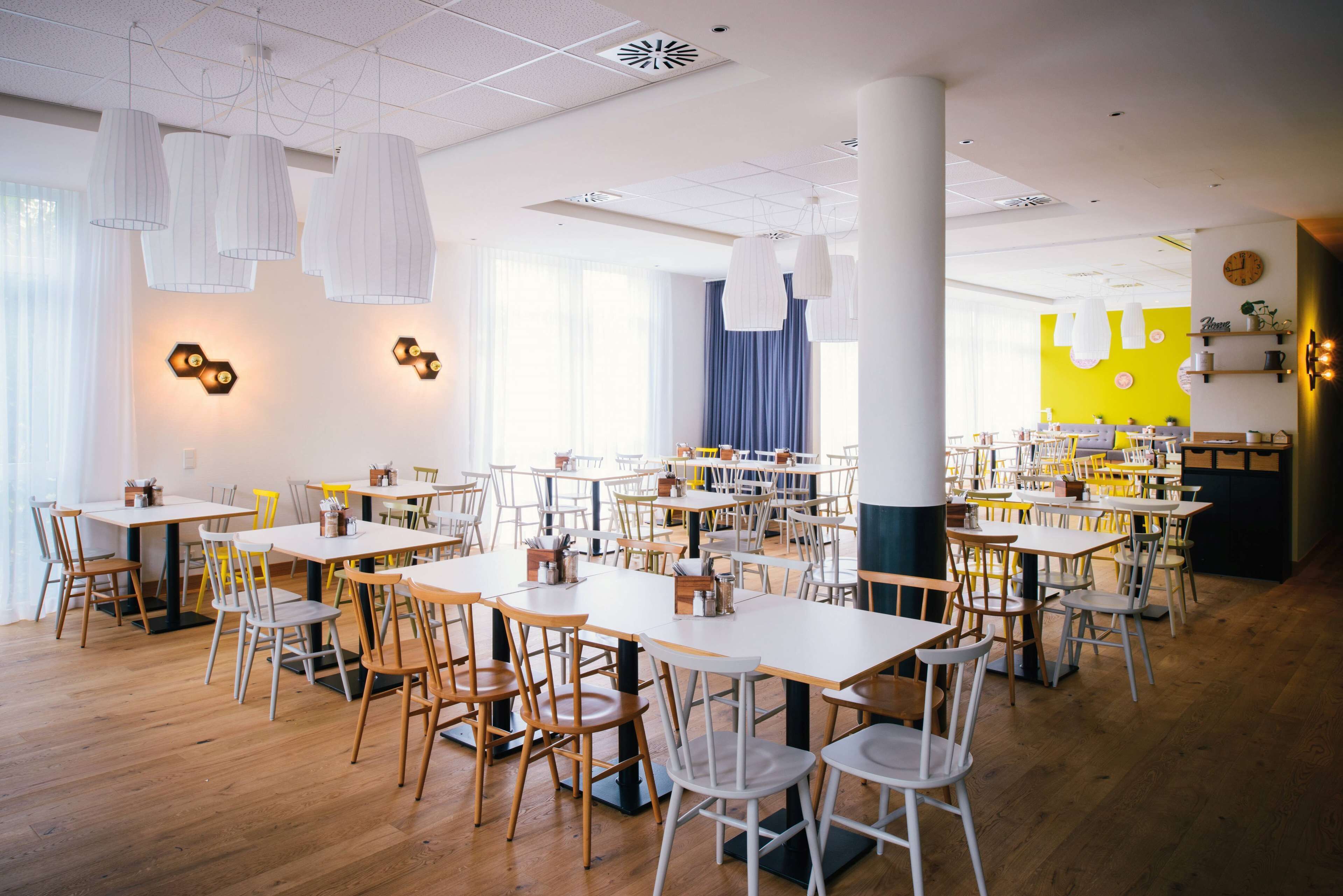 Een ruime eethoek met meerdere tafels en stoelen, een heldergele accentmuur en hanglampen.