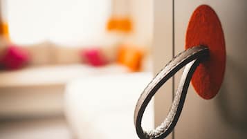 Uma maçaneta de metal presa a um objeto circular vermelho, provavelmente uma porta ou painel, em um quarto com fundo desfocado sugerindo um hotel ou estabelecimento similar.