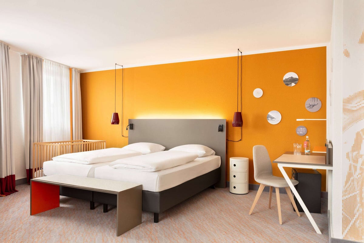 A modern hotel room with a bed, bedside table, chair, and a desk with a chair.