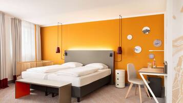 A modern hotel room with a bed, bedside table, chair, and a desk with a chair.