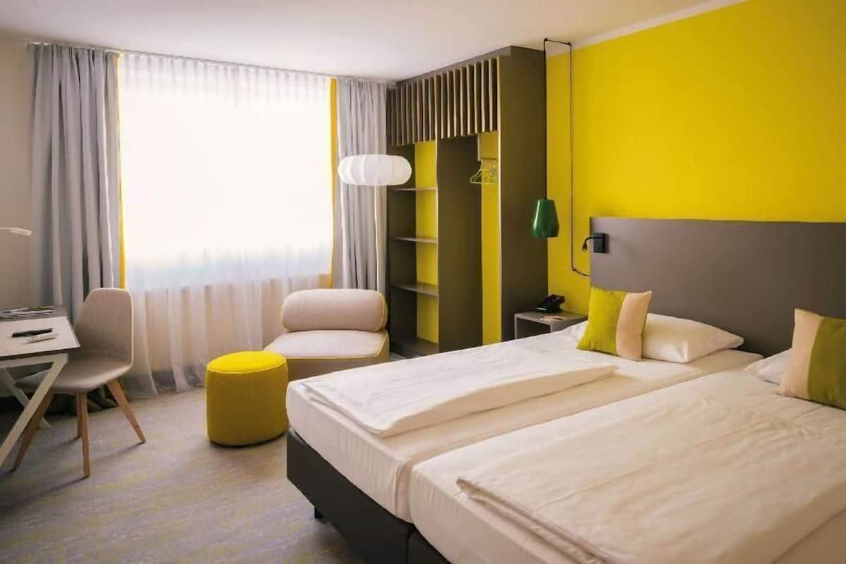 A hotel room with two beds, a desk, a chair, and a small ottoman.