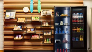 Máquina automática com bebidas e petiscos, e uma parede de madeira com diversos itens como livros, revistas e produtos embalados.