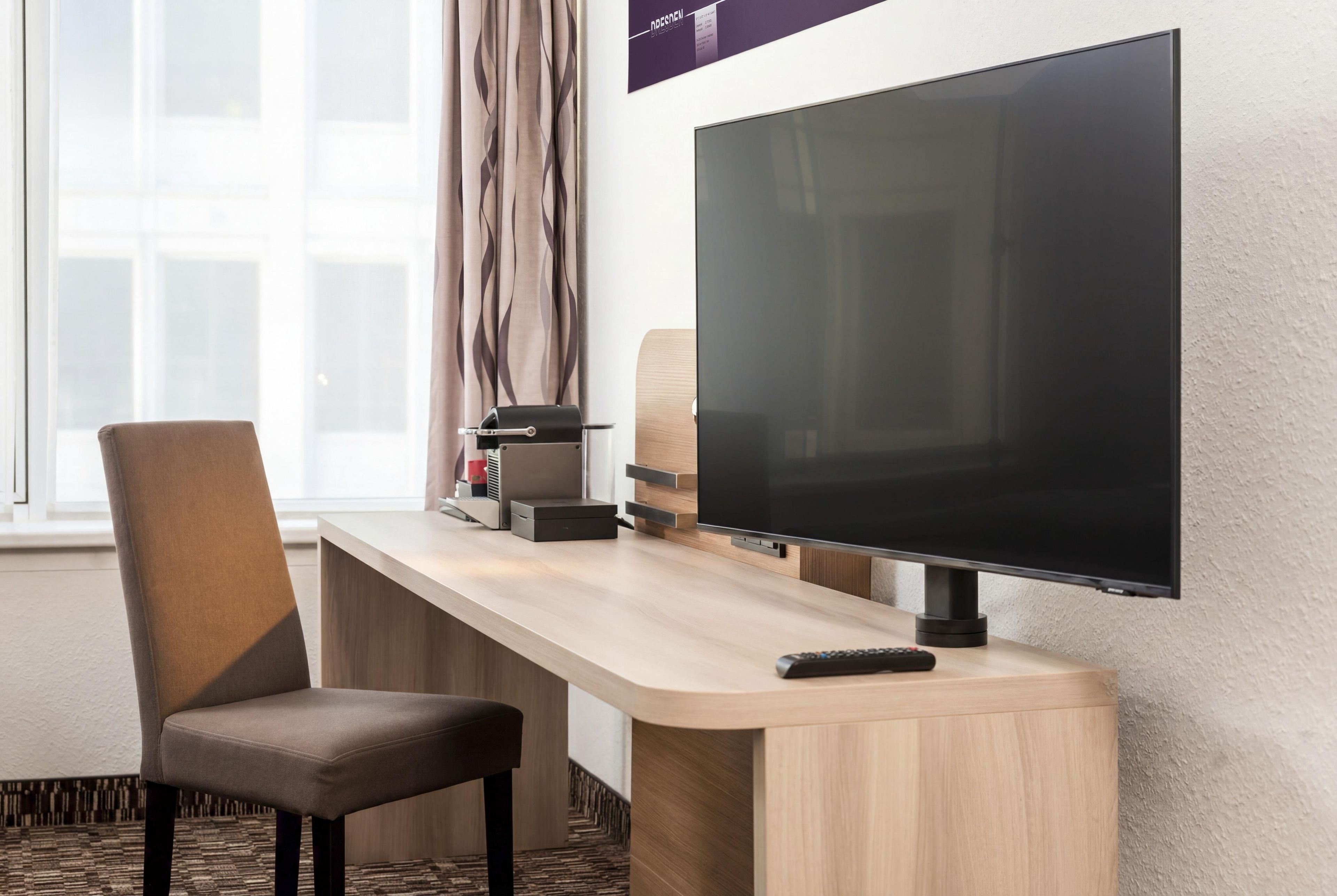 Ein Hotelzimmer mit Flachbildfernseher an der Wand, einem Holztisch mit Stuhl und einer Fernbedienung auf dem Tisch.