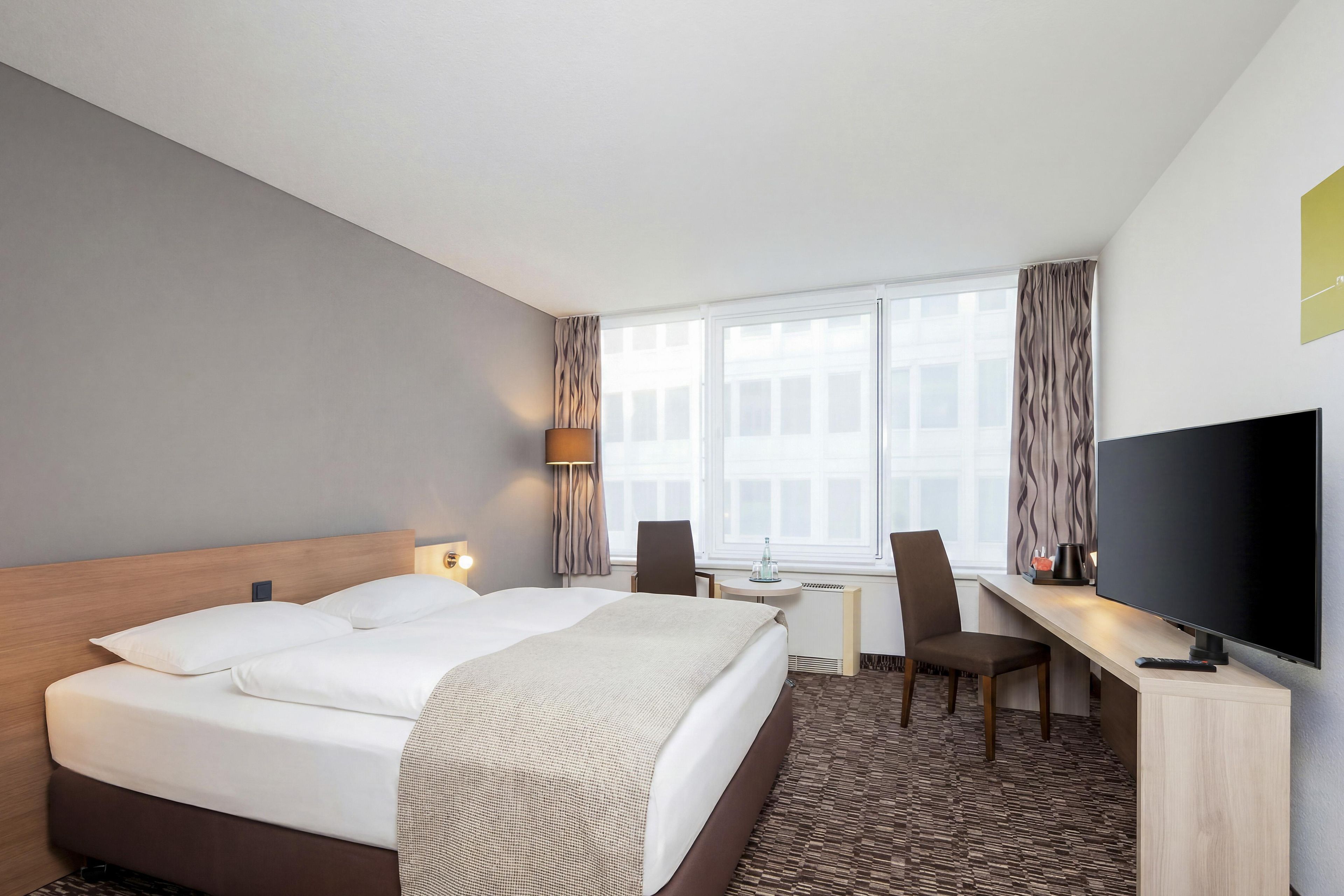 Ein Hotelzimmer mit einem großen Bett, einem Schreibtisch, einem Stuhl, einem Fernseher und einem Fenster mit Vorhängen.