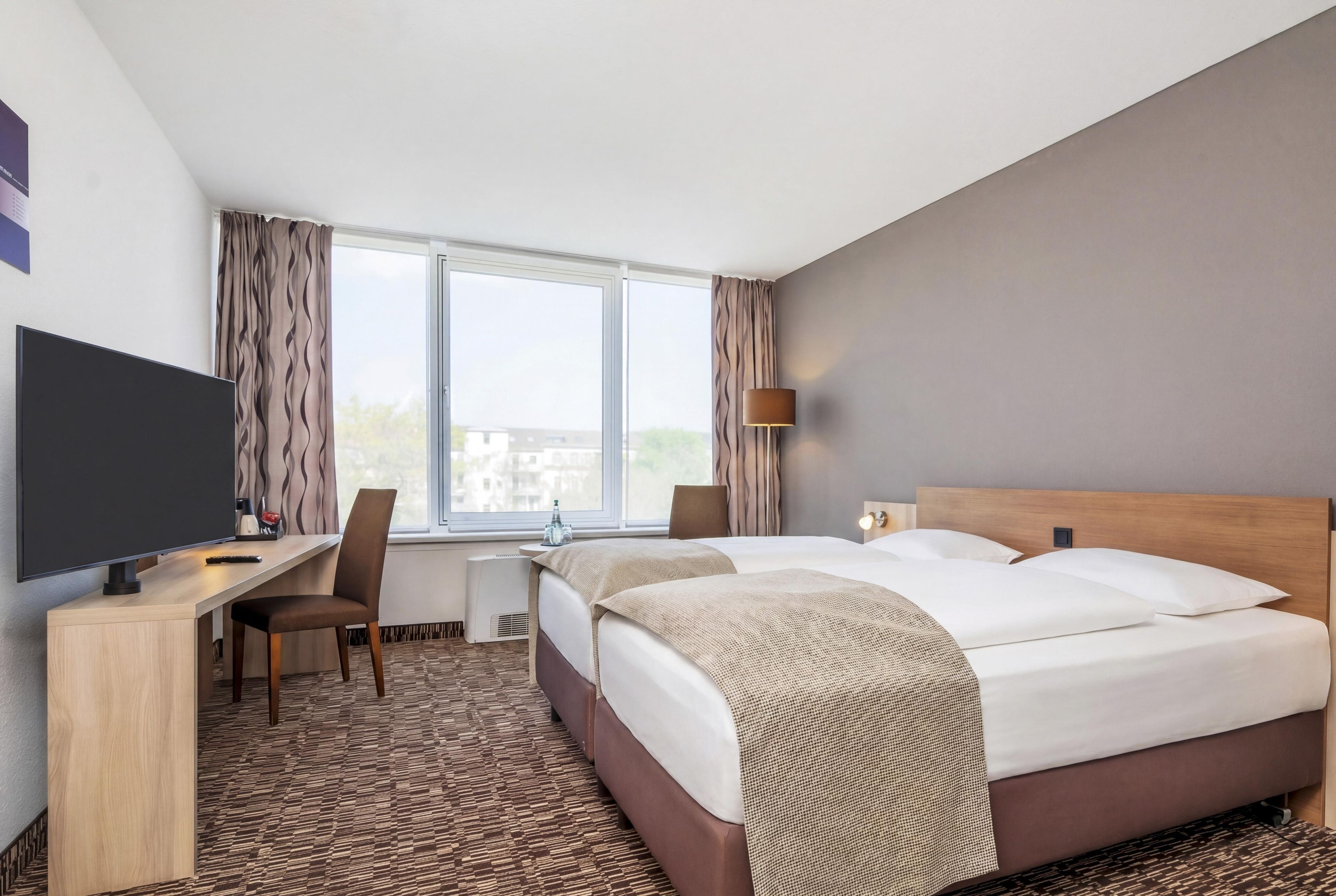Quarto de hotel com uma cama, escrivaninha, cadeira, televisão e janela com cortinas.