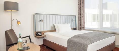 A hotel room with a bed, a bedside table with a lamp, a chair, and a view of the outside.