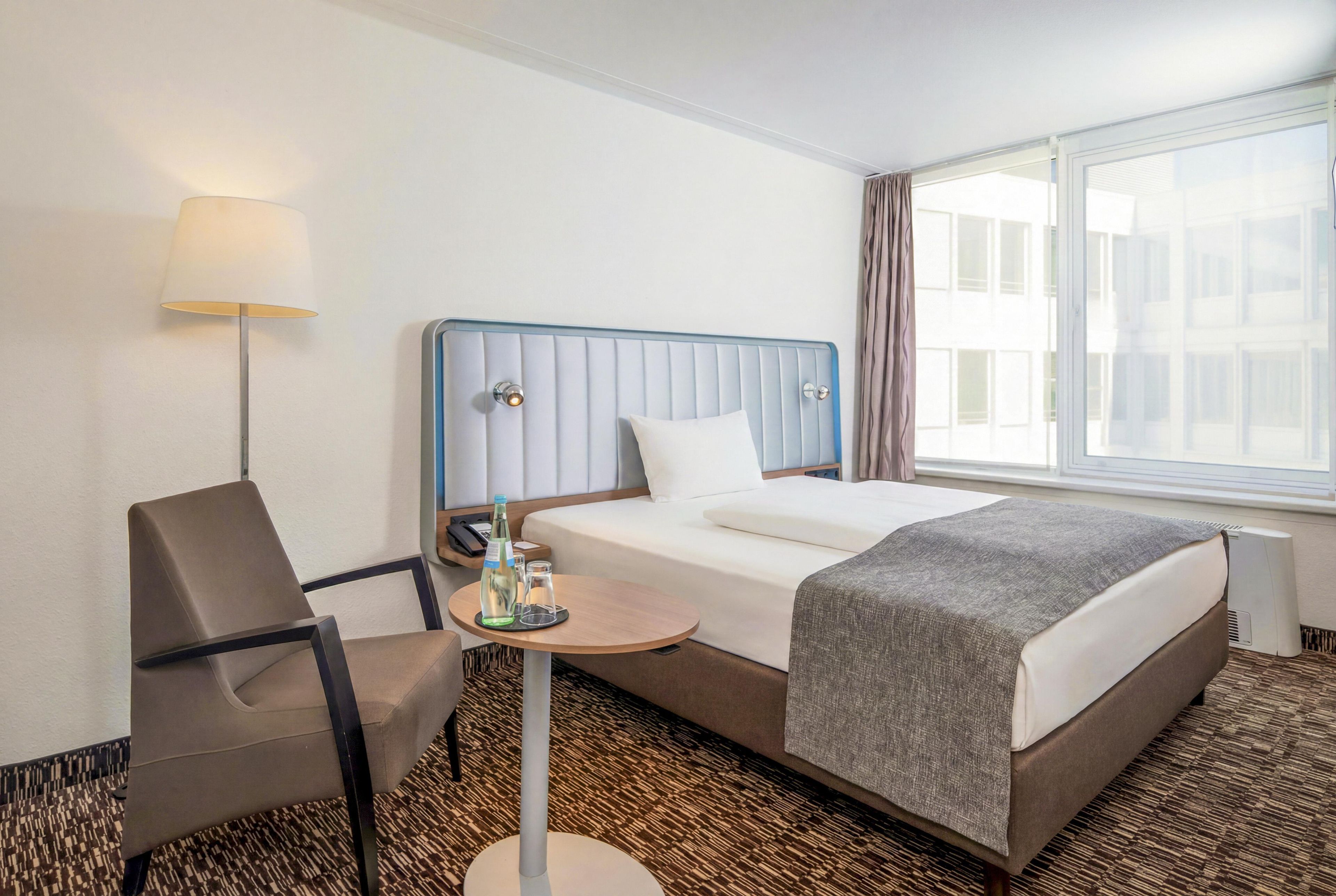 Habitación de hotel con cama, una silla, una mesita, lámpara y ventana con vistas a edificios.