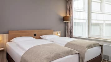 Ein Hotelzimmer mit Doppelbett, Nachttisch, Lampe, einem Fenster mit Vorhängen und einer Wandleuchte.
