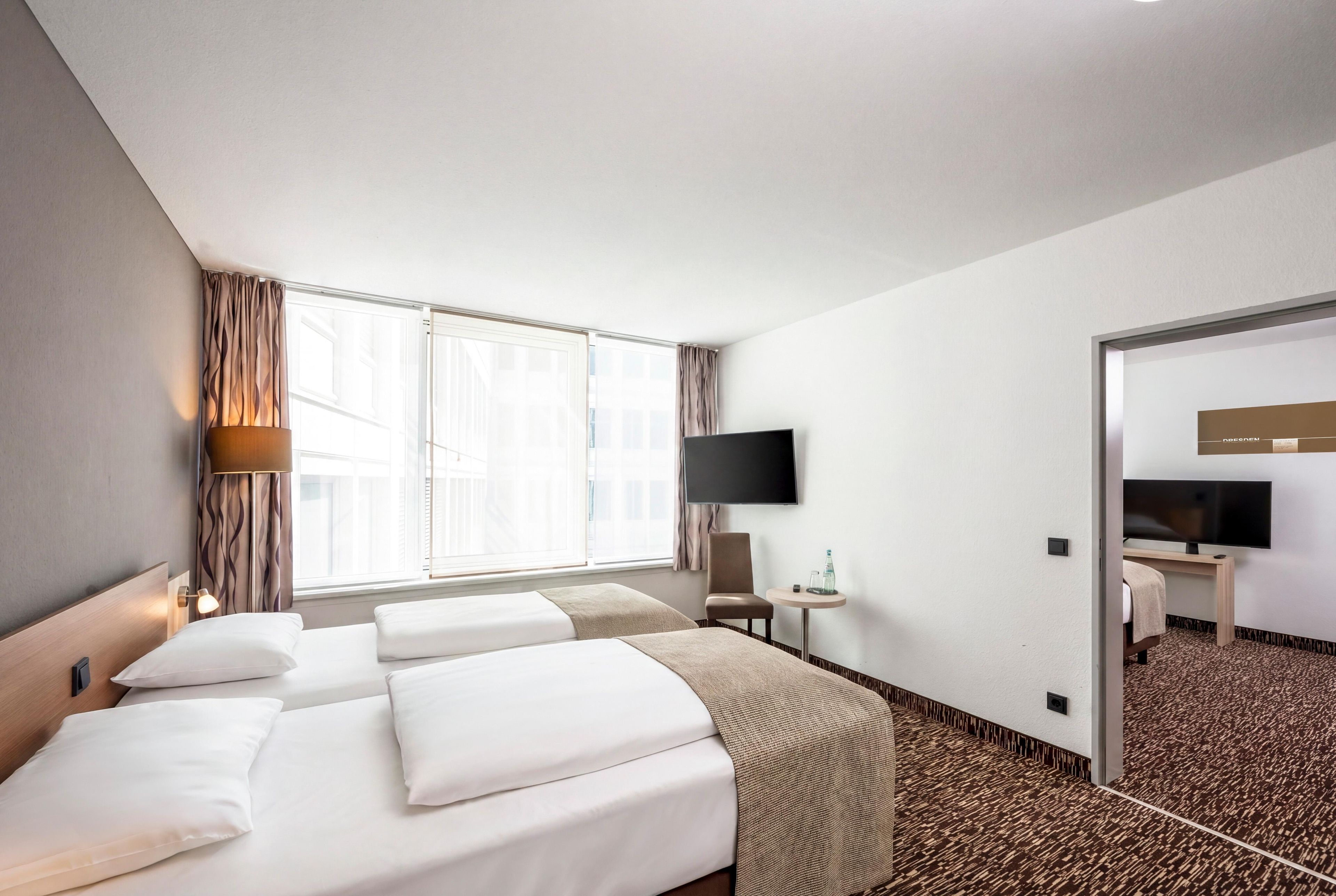 Ein Hotelzimmer mit einem großen Bett, einem Flachbildfernseher, einem kleinen Tisch mit einer Flasche und Blick auf die Stadt durch das Fenster.