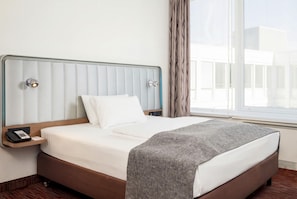 Une chambre d’hôtel avec un lit, une table de chevet avec un téléphone, une fenêtre avec des rideaux et une vue sur les bâtiments environnants.