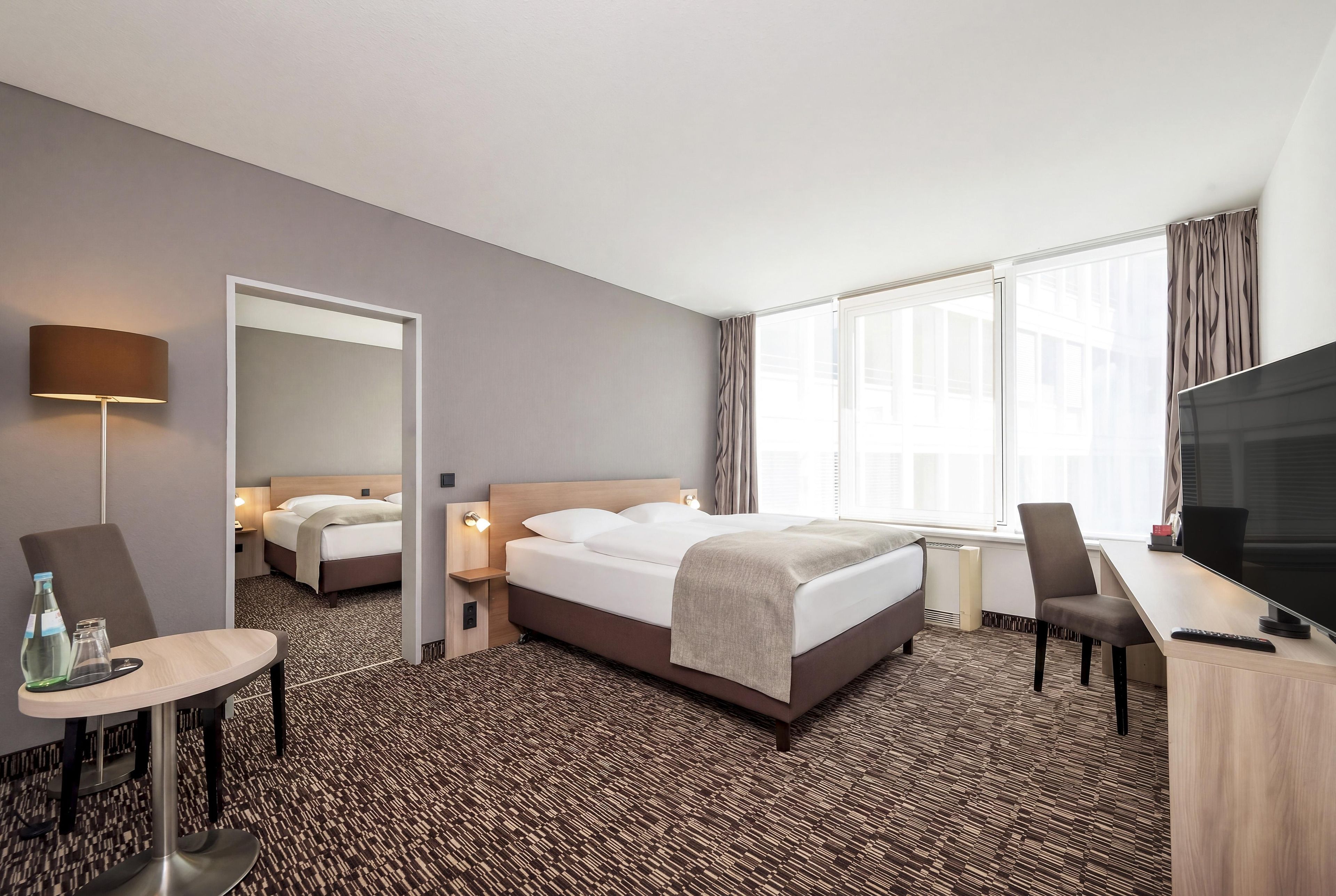 A hotel room with a bed, a desk, a chair, a TV, and a window with curtains.