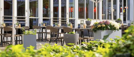 An outdoor seating area with tables and chairs, potted plants, and a glass enclosure.