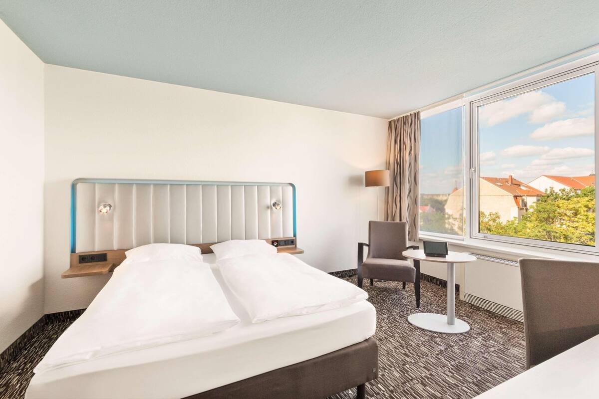 Hotelkamer met een bed, een stoel, een tafeltje en een raam met uitzicht op gebouwen.