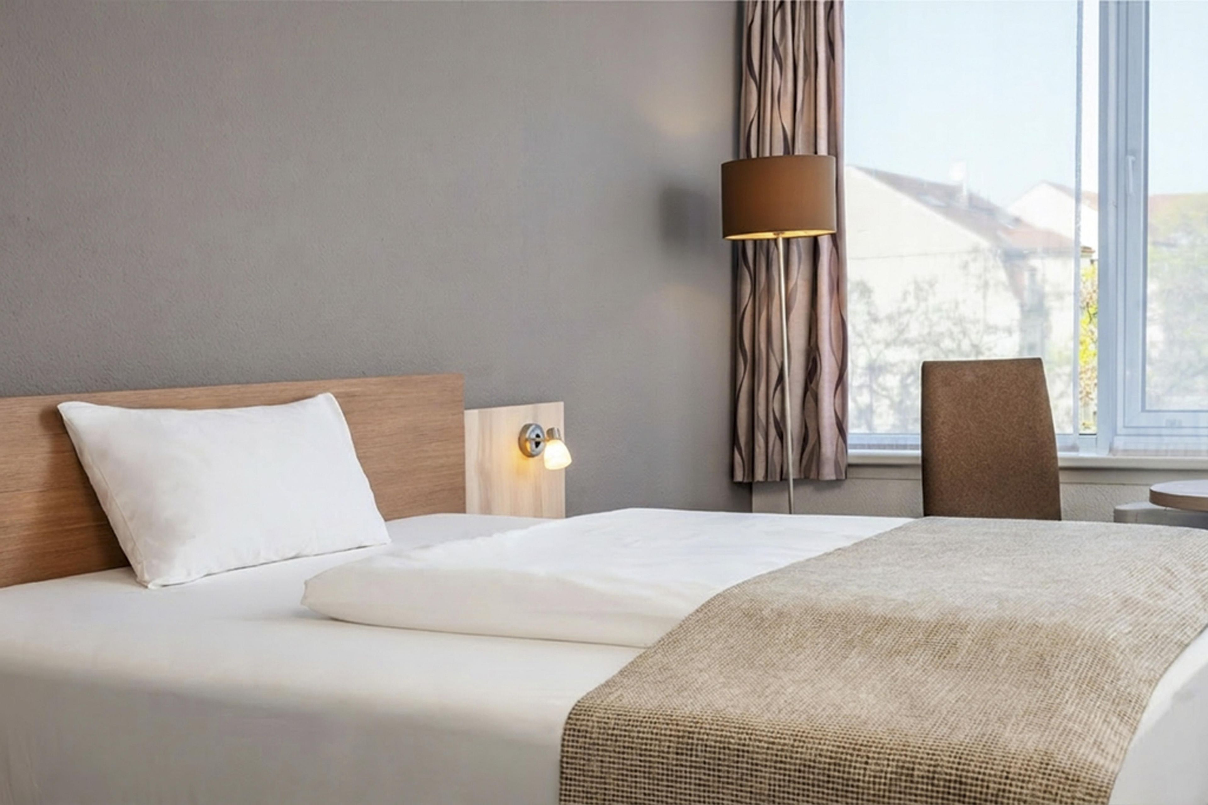 Ein Hotelzimmer mit einem Bett, einer Nachttischlampe, einem Sessel und einem Fenster mit Vorhängen.