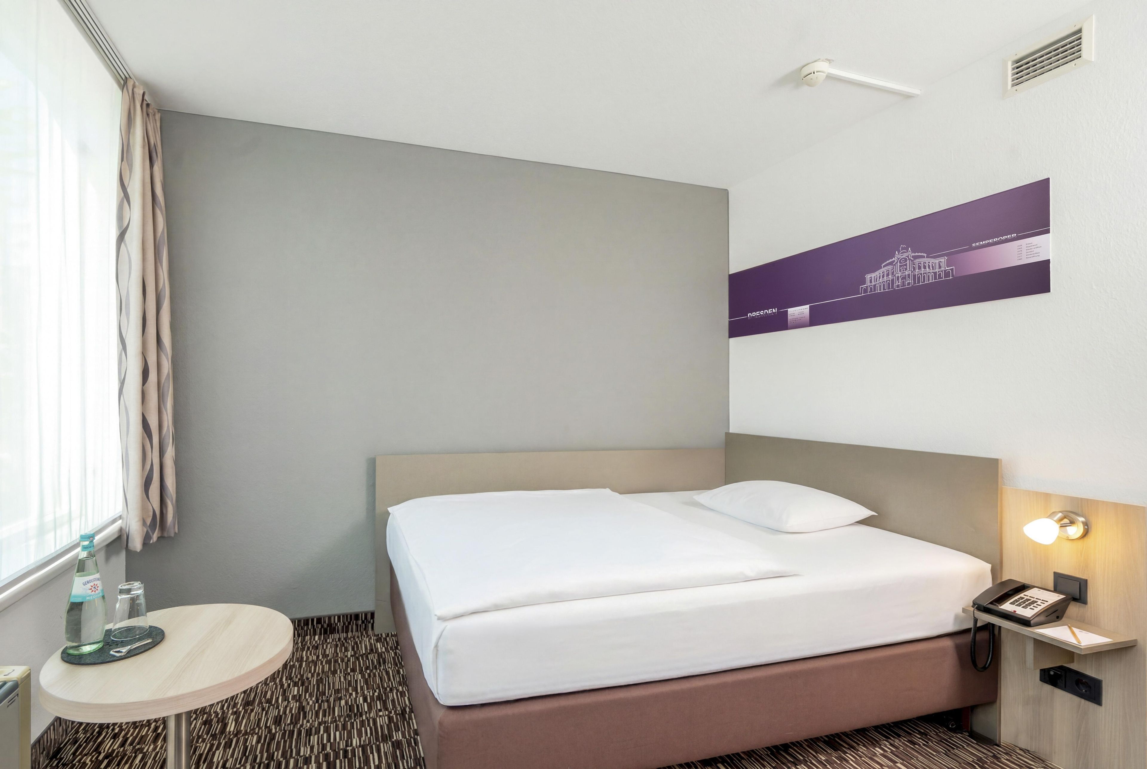 Ein Hotelzimmer mit einem Bett, einem Nachttisch mit Lampe, einem an der Wand montierten Telefon und einem Fenster mit Vorhängen.
