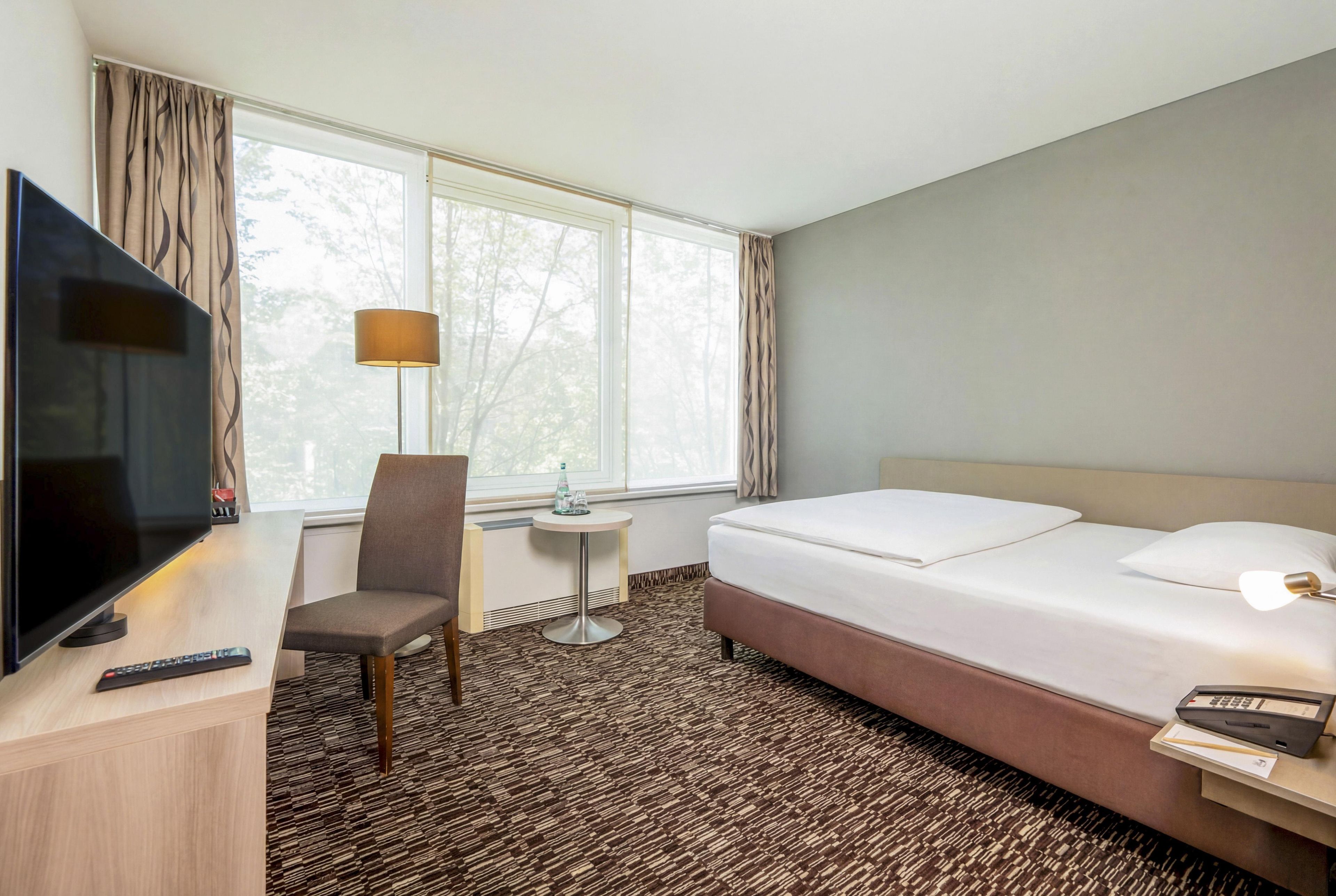 A hotel room with a bed, a desk with a TV, a chair, a lamp, and a window with curtains.