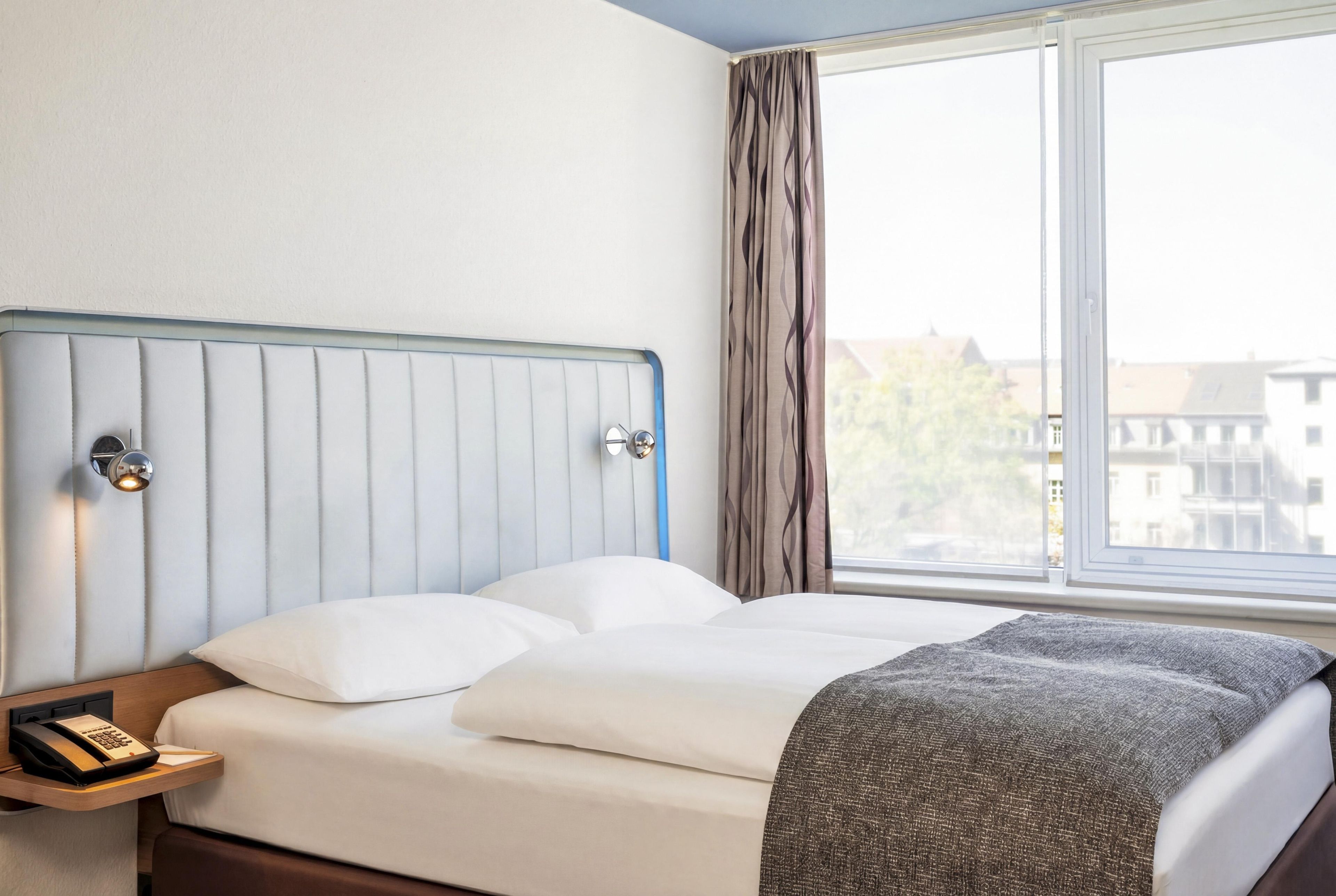 Ein Hotelzimmer mit einem Bett, einem Nachttischtelefon, einem Fenster mit Vorhängen und Blick auf Gebäude.