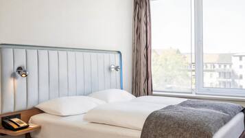 Ein Hotelzimmer mit einem Bett, einem Nachttischtelefon, einem Fenster mit Vorhängen und Blick auf Gebäude.