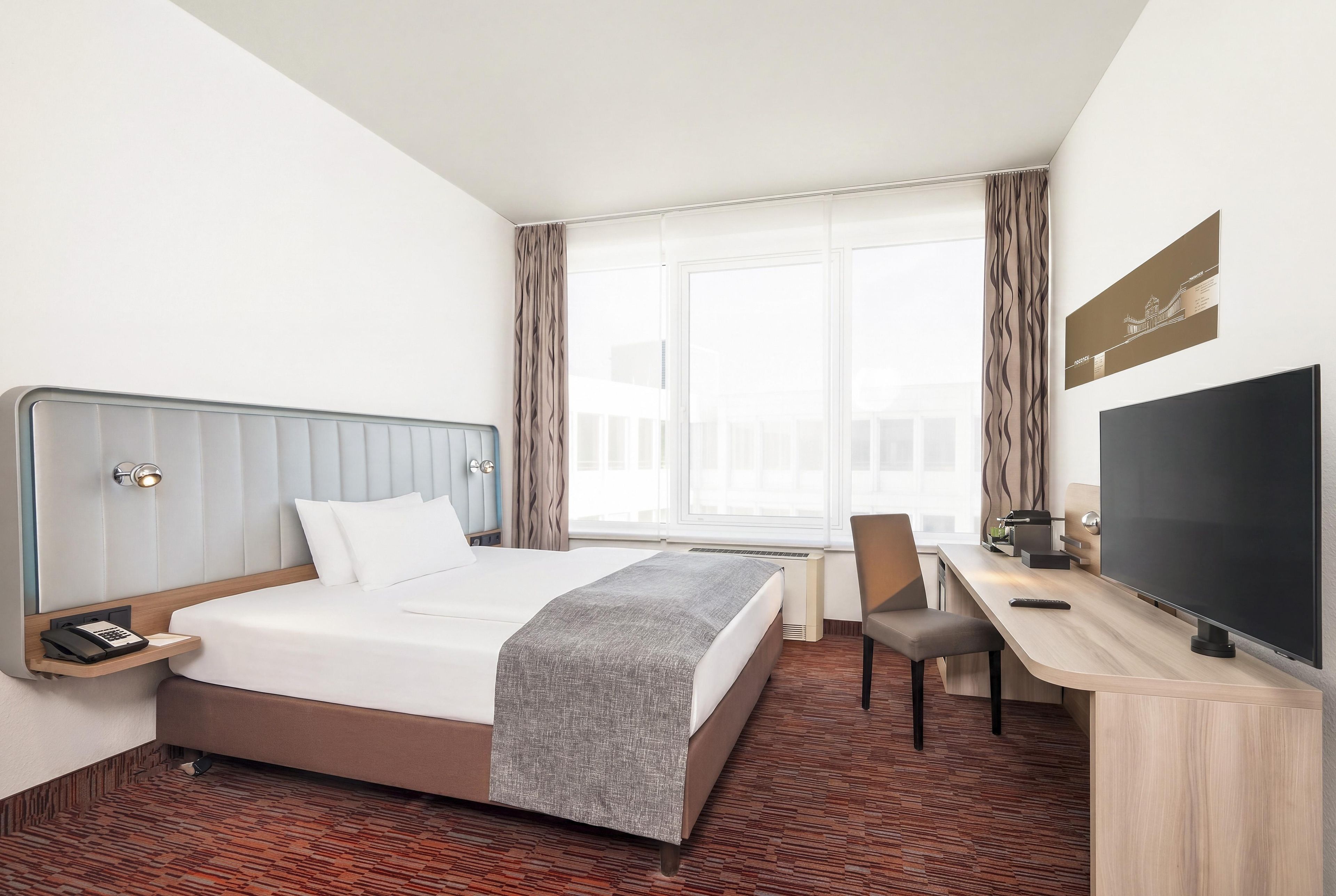 Ein Hotelzimmer mit einem Bett, einem Schreibtisch, einem Sessel, einem Fernseher und einem fenster mit Vorhängen.