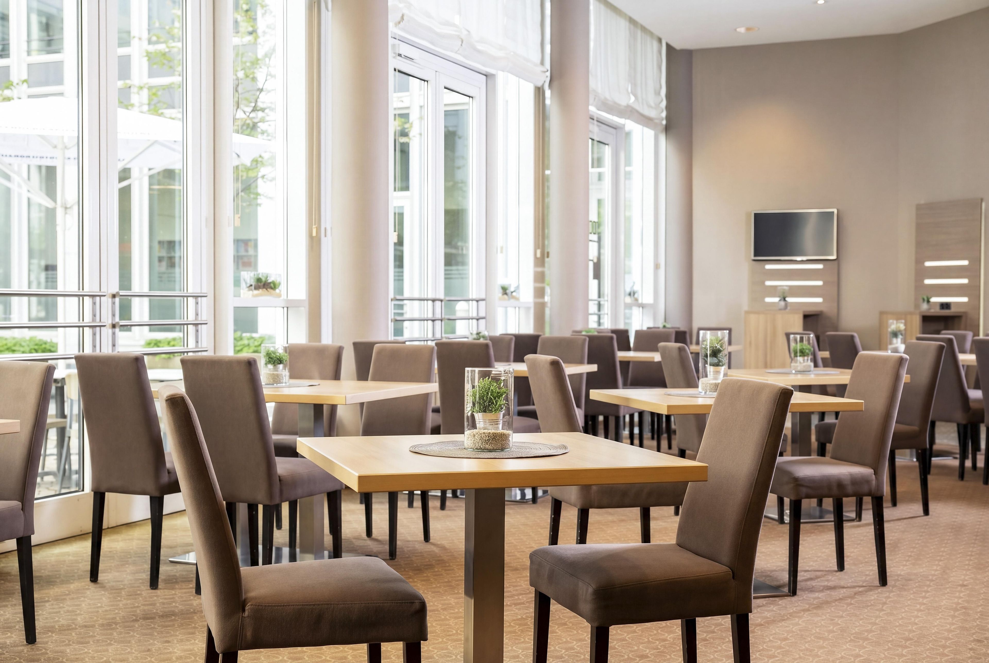 Un espace repas moderne doté de plusieurs tables et chaises, de grandes fenêtres et d’un téléviseur à écran plat fixé au mur.