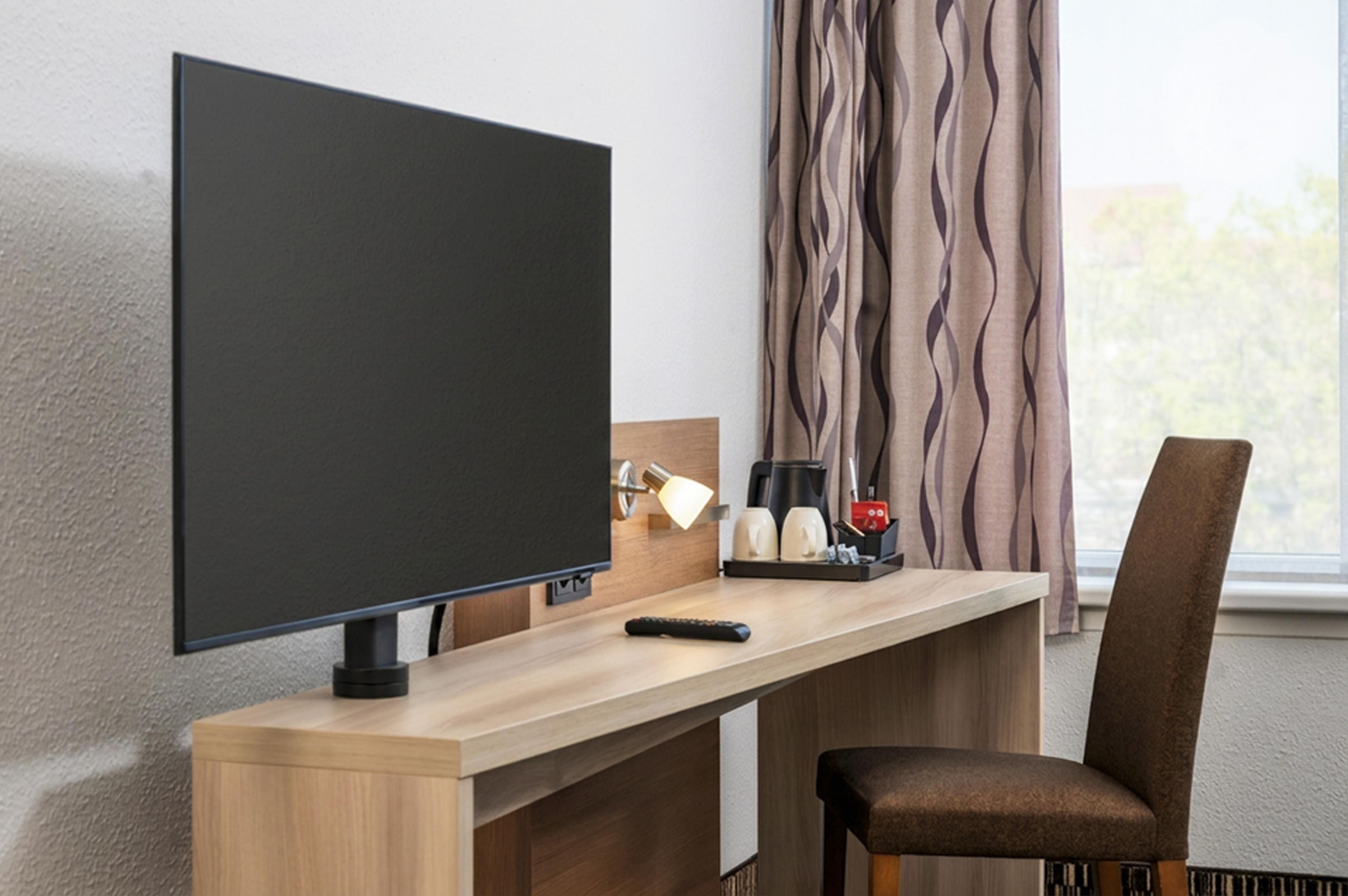 Ein Hotelzimmer mit einem Flachbildfernseher an einer Holztischplatte, einer Fernbedienung, einer Kaffeemaschine und einem Sessel.