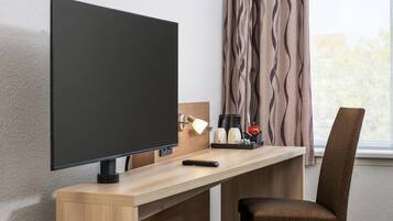 Ein Hotelzimmer mit einem Flachbildfernseher an einer Holztischplatte, einer Fernbedienung, einer Kaffeemaschine und einem Sessel.