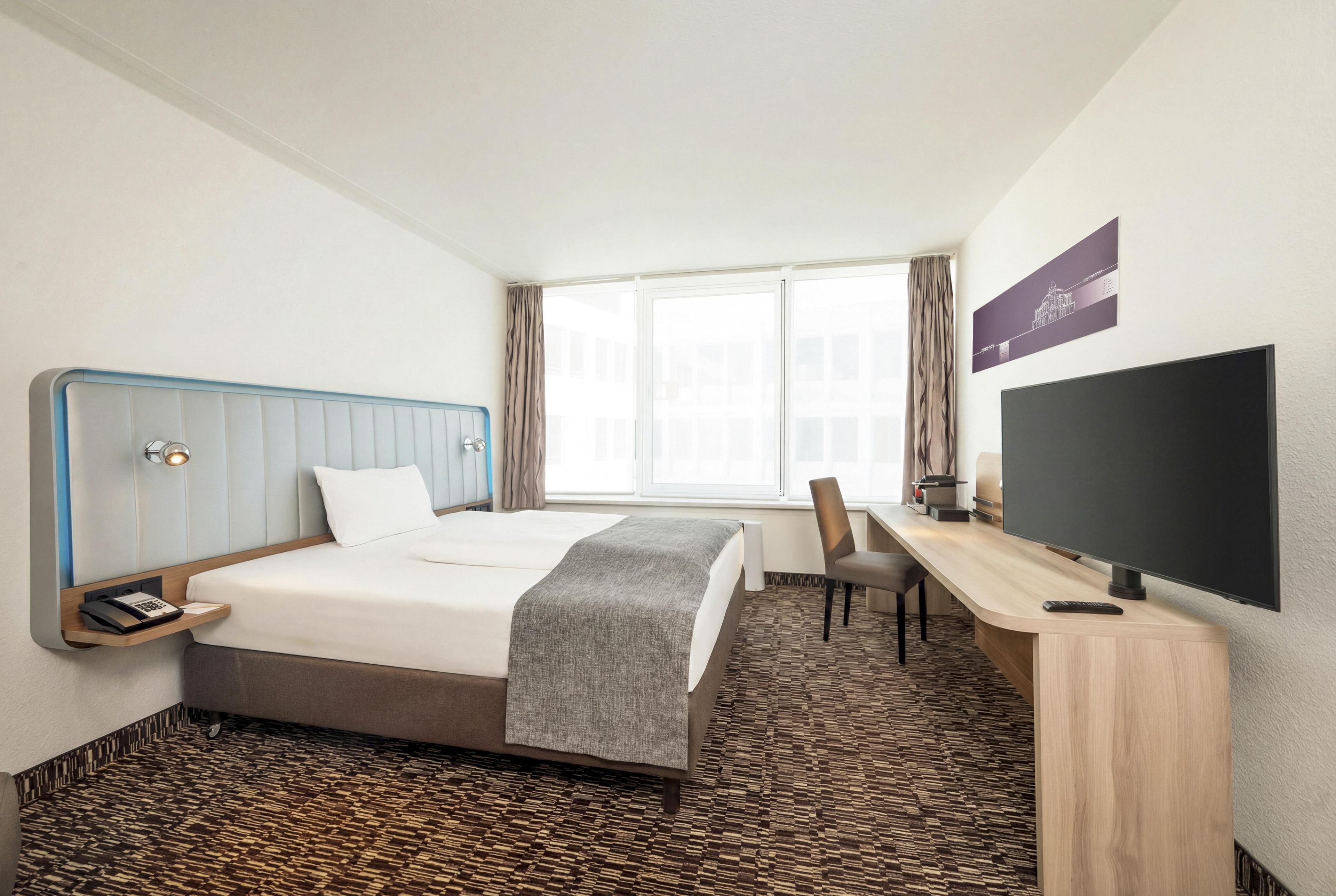 Ein Hotelzimmer mit Bett, Schreibtisch, Stuhl, Fernseher und einem Fenster mit Vorhängen.