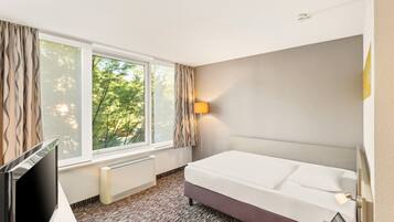 Ein Hotelzimmer mit Bett, Fernseher, Fenster mit Vorhängen, einer Wandlampe und einem Heizkörper.