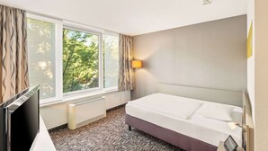 A hotel room with a bed, a TV, a window with curtains, a lamp, and a view of trees.