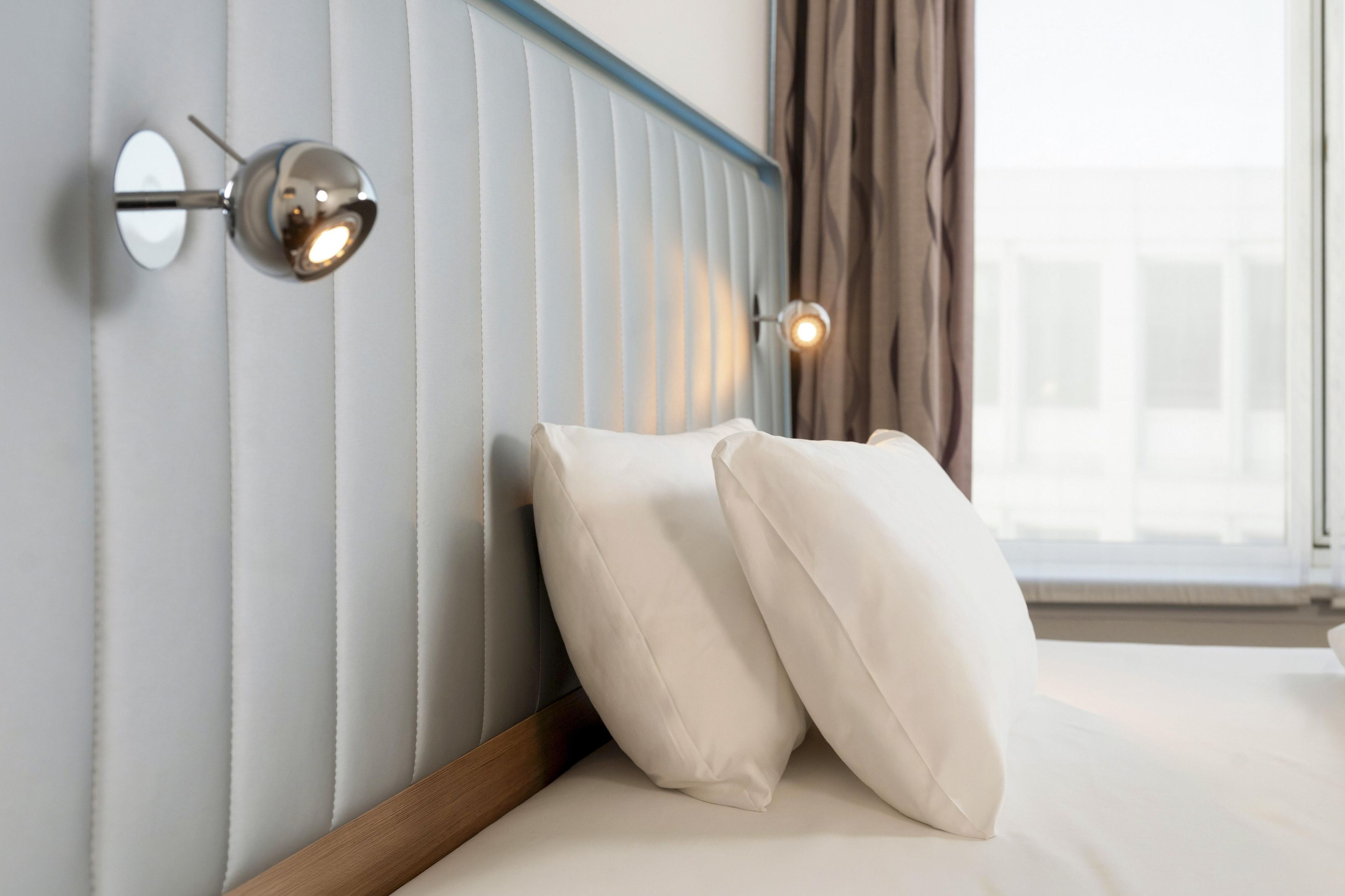A hotel room with a bed, white pillows, a headboard with vertical lines, and wall-mounted lights.