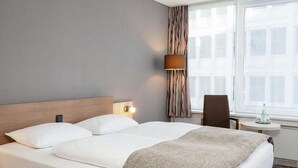 Habitación de hotel con cama, lámpara de noche, una silla, una mesita y ventana con cortinas.