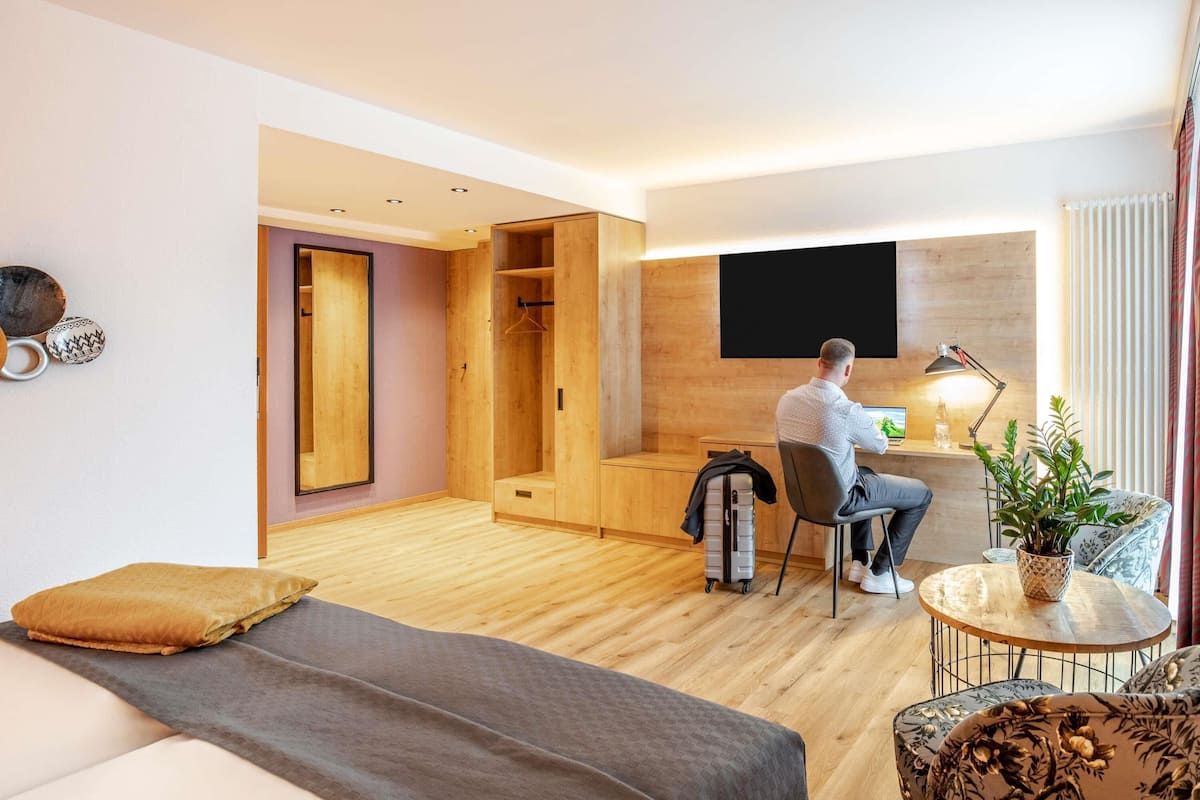 A modern hotel room with a wooden floor, a bed, a desk with a chair, a TV mounted on the wall, and a small table with a plant.