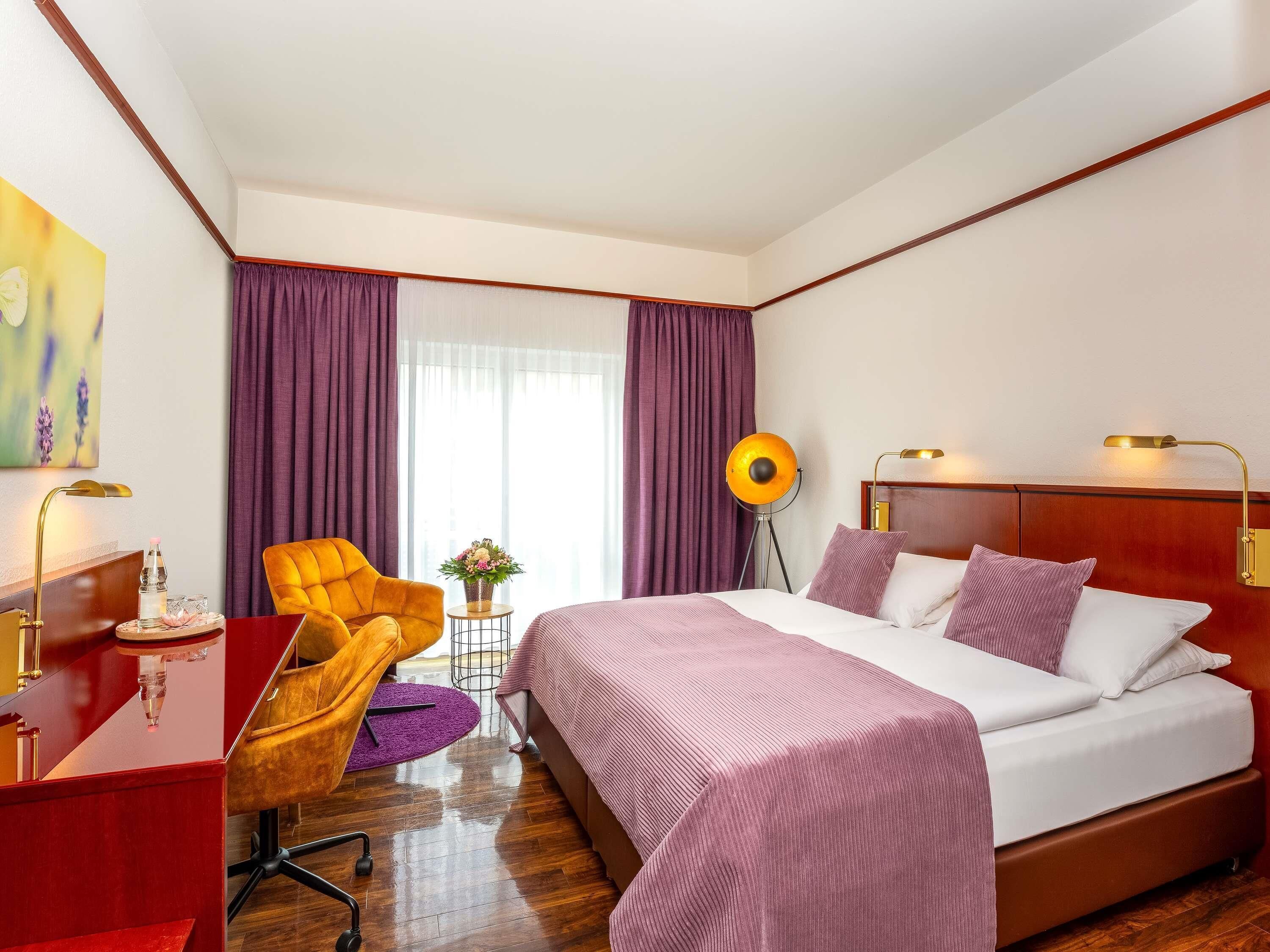 Une chambre d’hôtel avec un lit, un bureau, une chaise, une lampe et une fenêtre avec des rideaux.