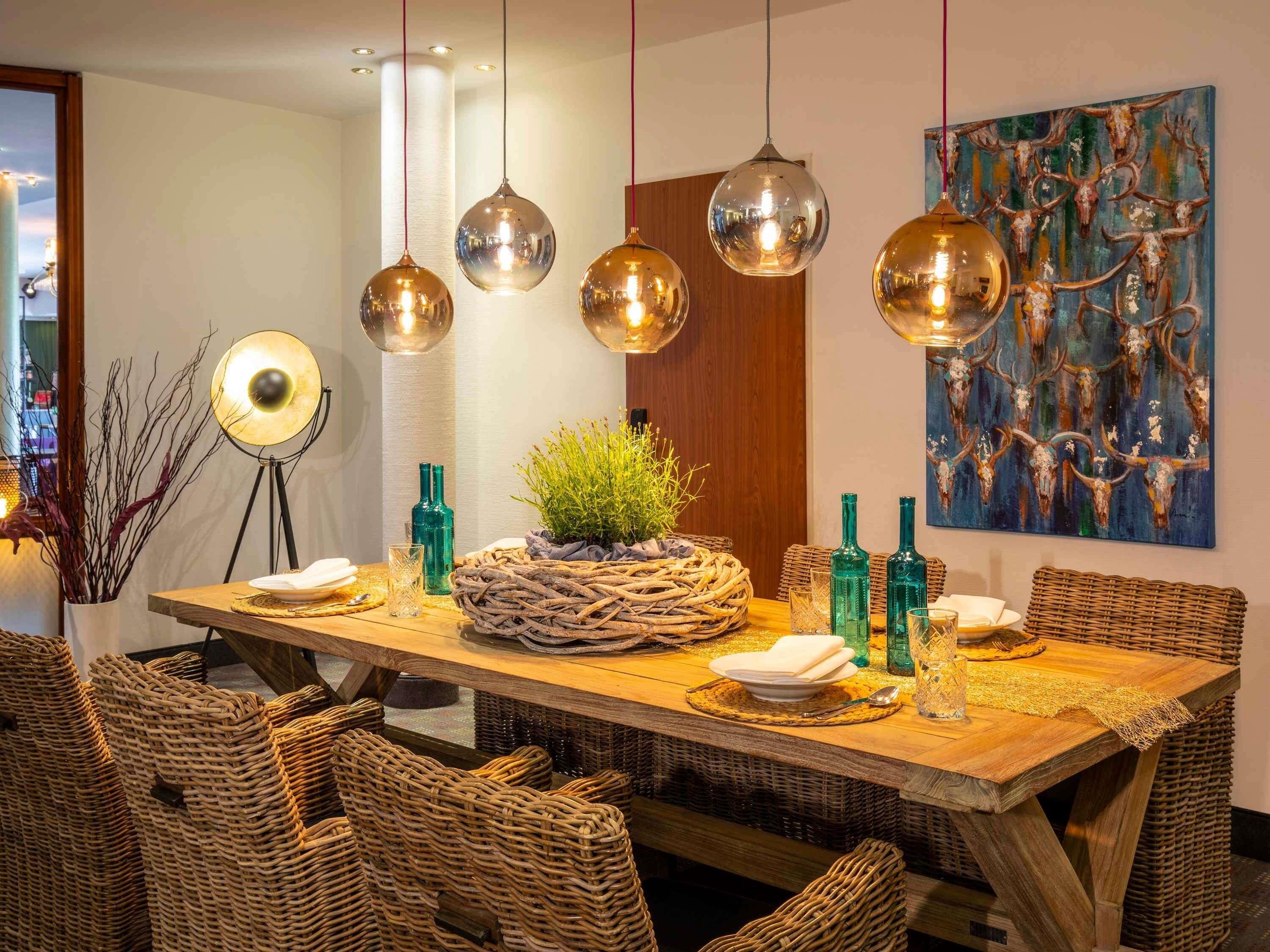 A dining area with a wooden table, wicker chairs, hanging pendant lights, and a painting on the wall.