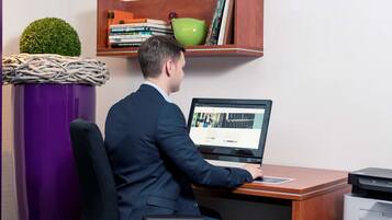 A man in a suit working at a desk with a laptop, a printer, and a bookshelf with books and a decorative bowl.