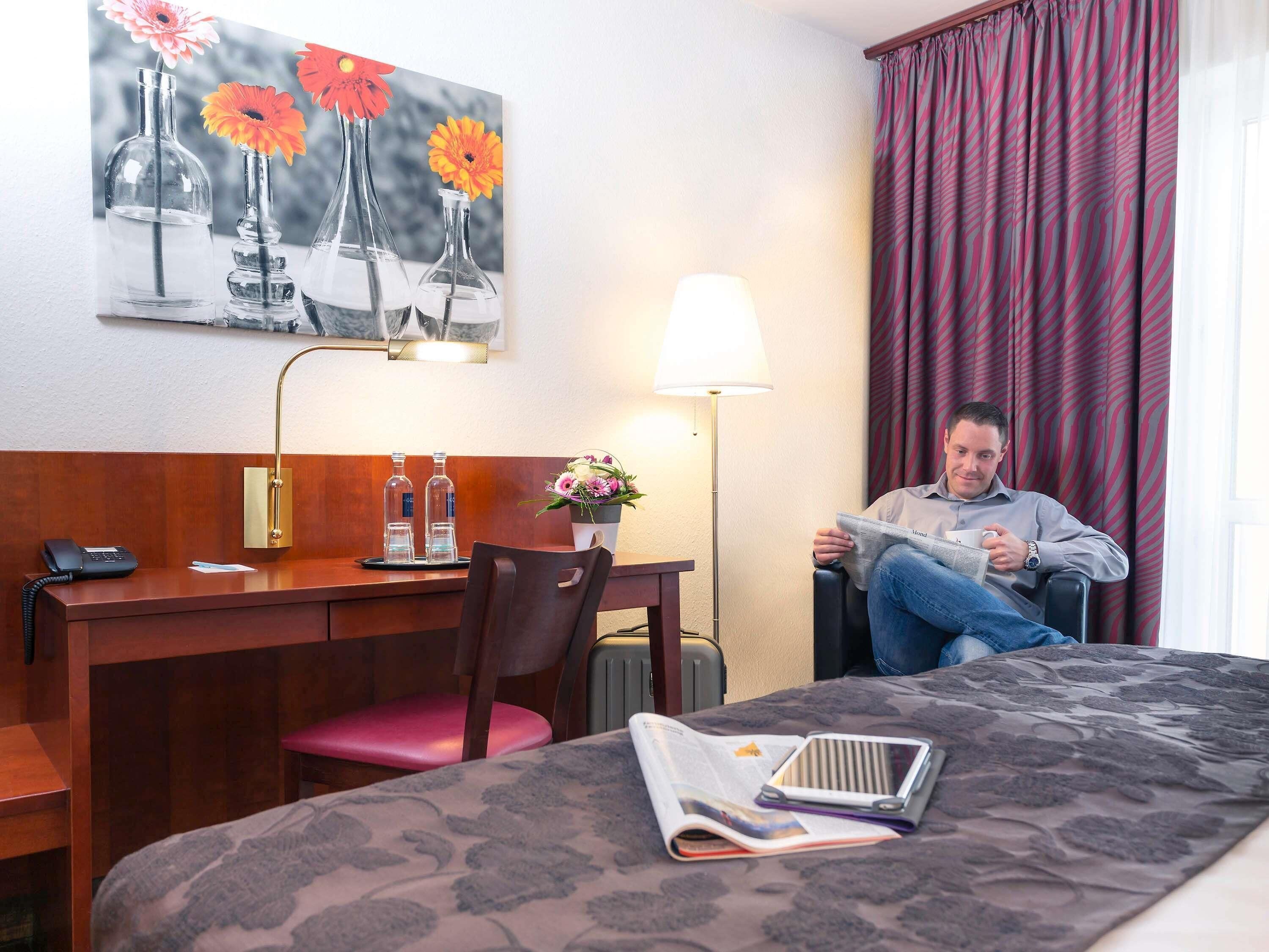 A hotel room with a bed, a desk with a telephone, a chair, a lamp, and a painting on the wall.