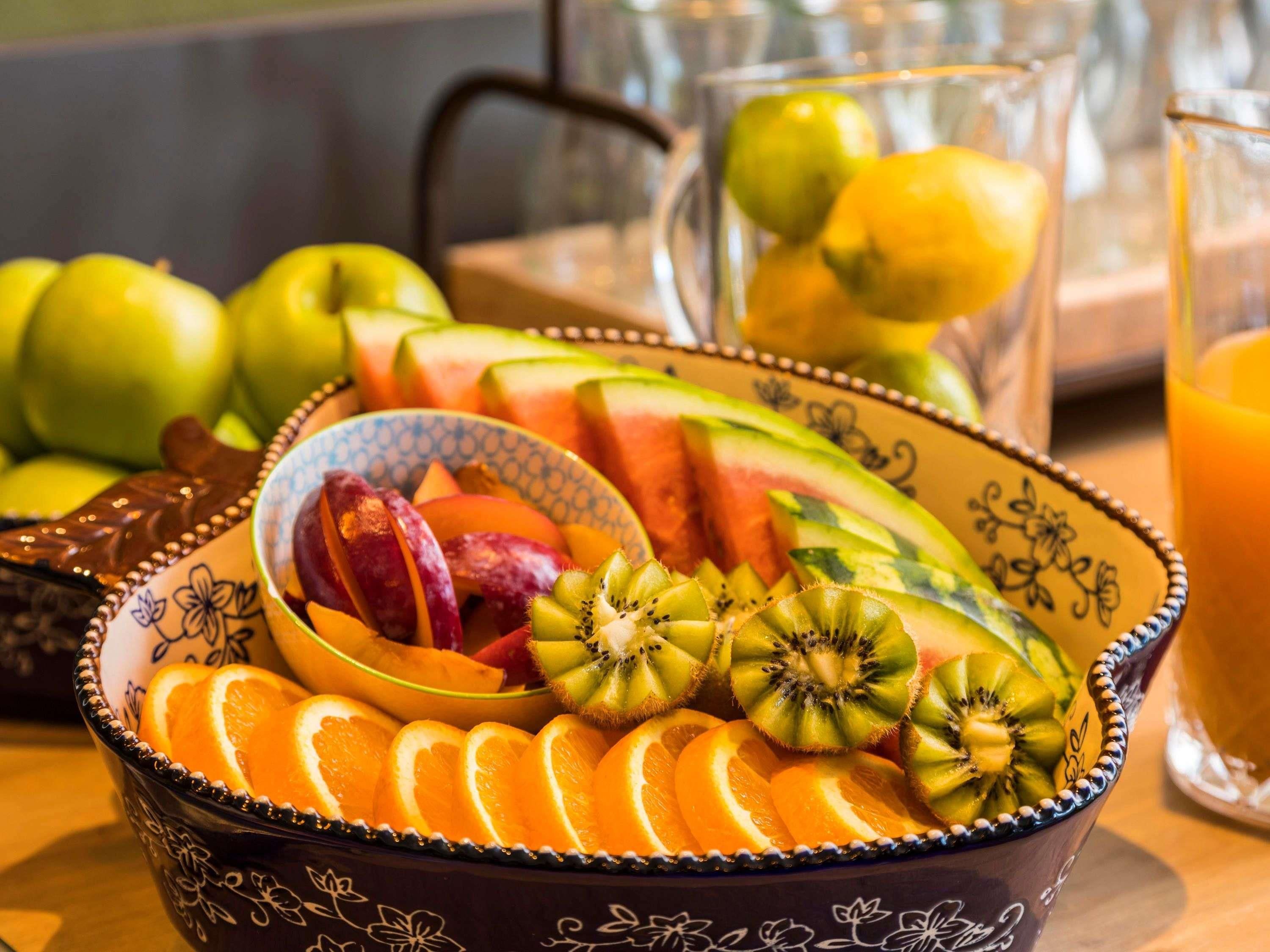 A bowl of sliced fruits including oranges, kiwi, and watermelon, with a glass pitcher and apples in the background.