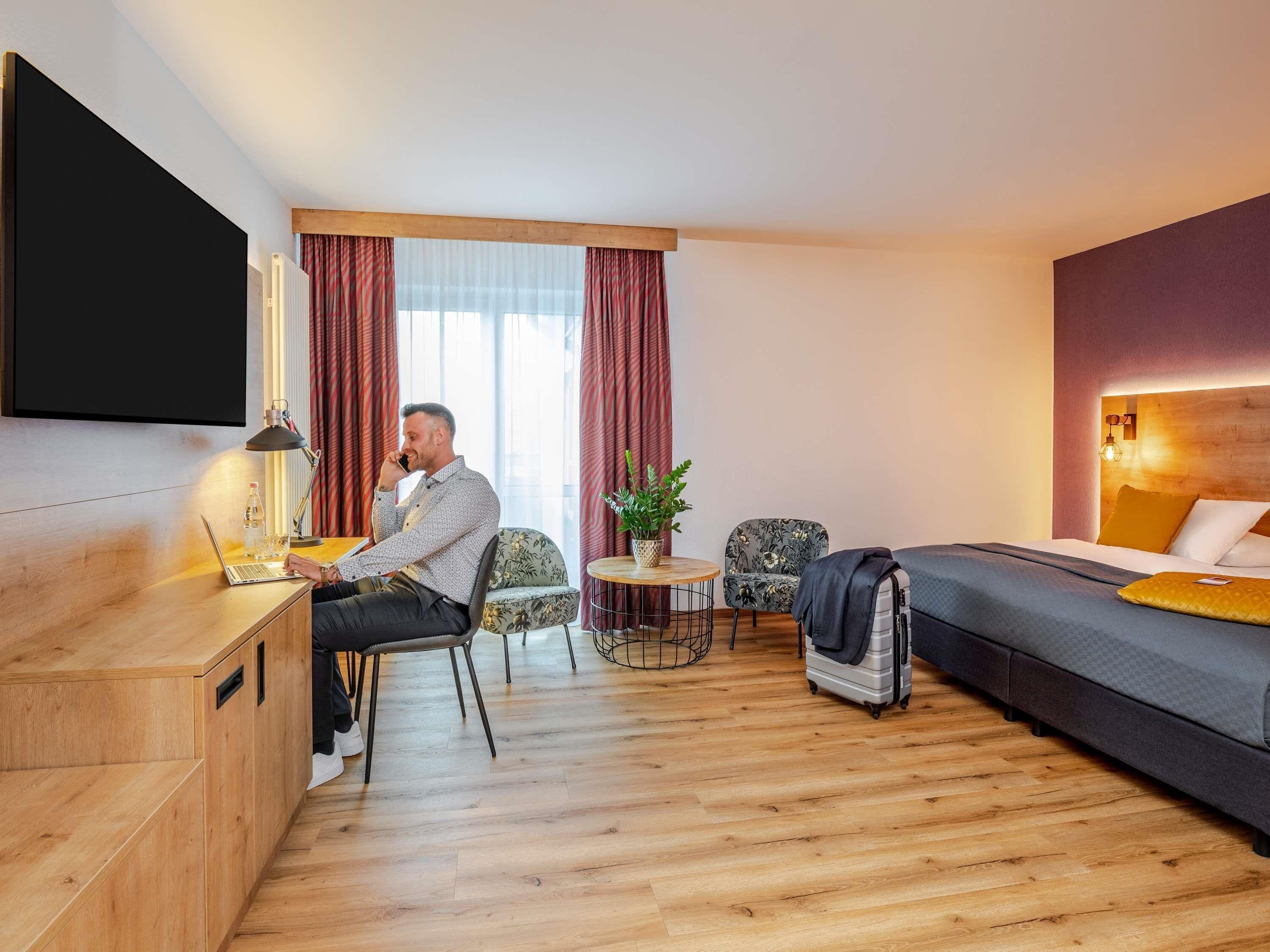 A hotel room with a desk, a bed, a TV, a window with curtains, a small table with a plant, and a chair.