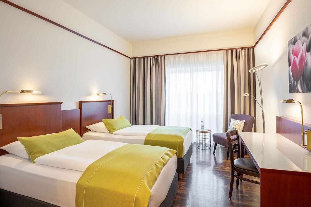 A hotel room with two beds, a desk, and a chair. There is a window with curtains, a lamp, and a picture on the wall.