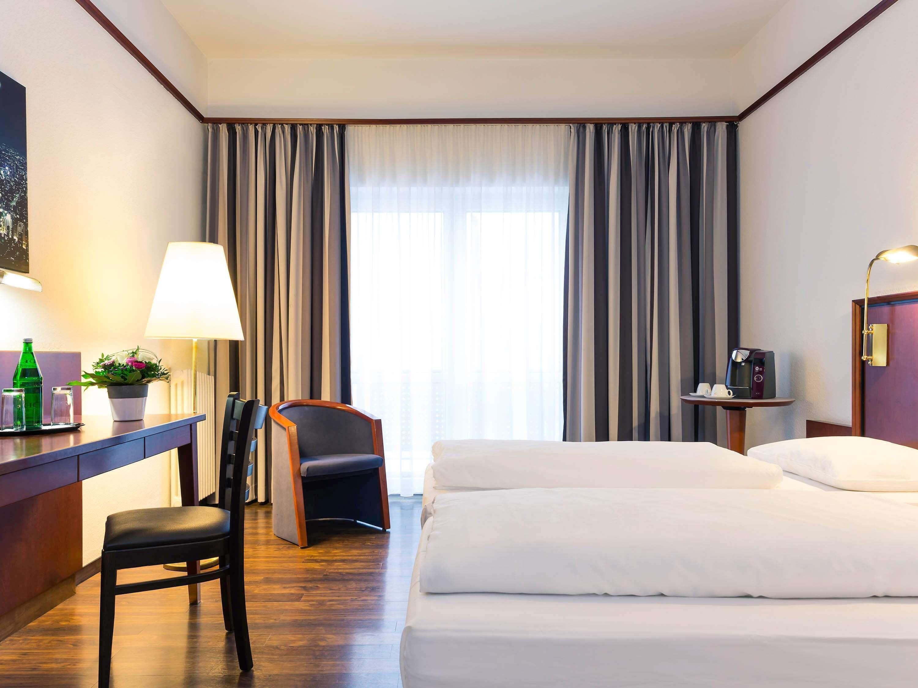 A hotel room with a bed, a desk with a lamp, a chair, a small table with a coffee maker, and a window with curtains.
