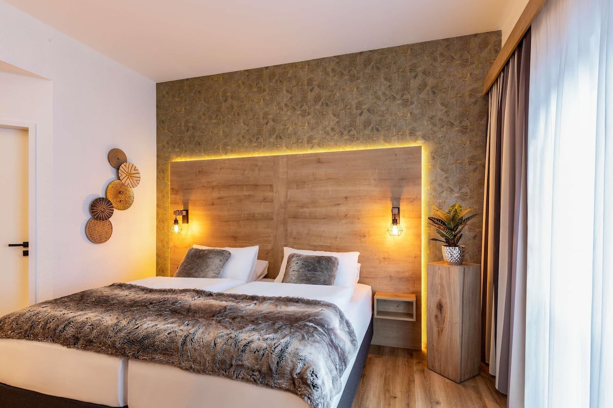 A modern bedroom with a large bed, wooden headboard, wall-mounted lights, and a small plant.