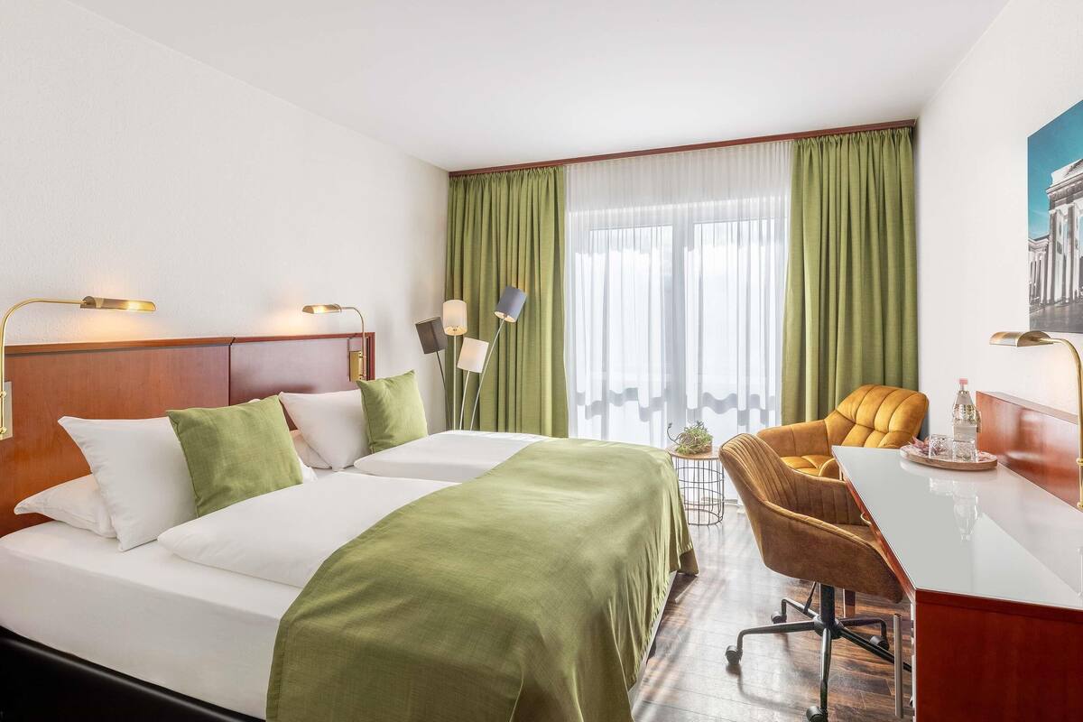 A hotel room with a bed, two bedside lamps, a desk with a chair, a small table with a plant, and a window with green curtains.