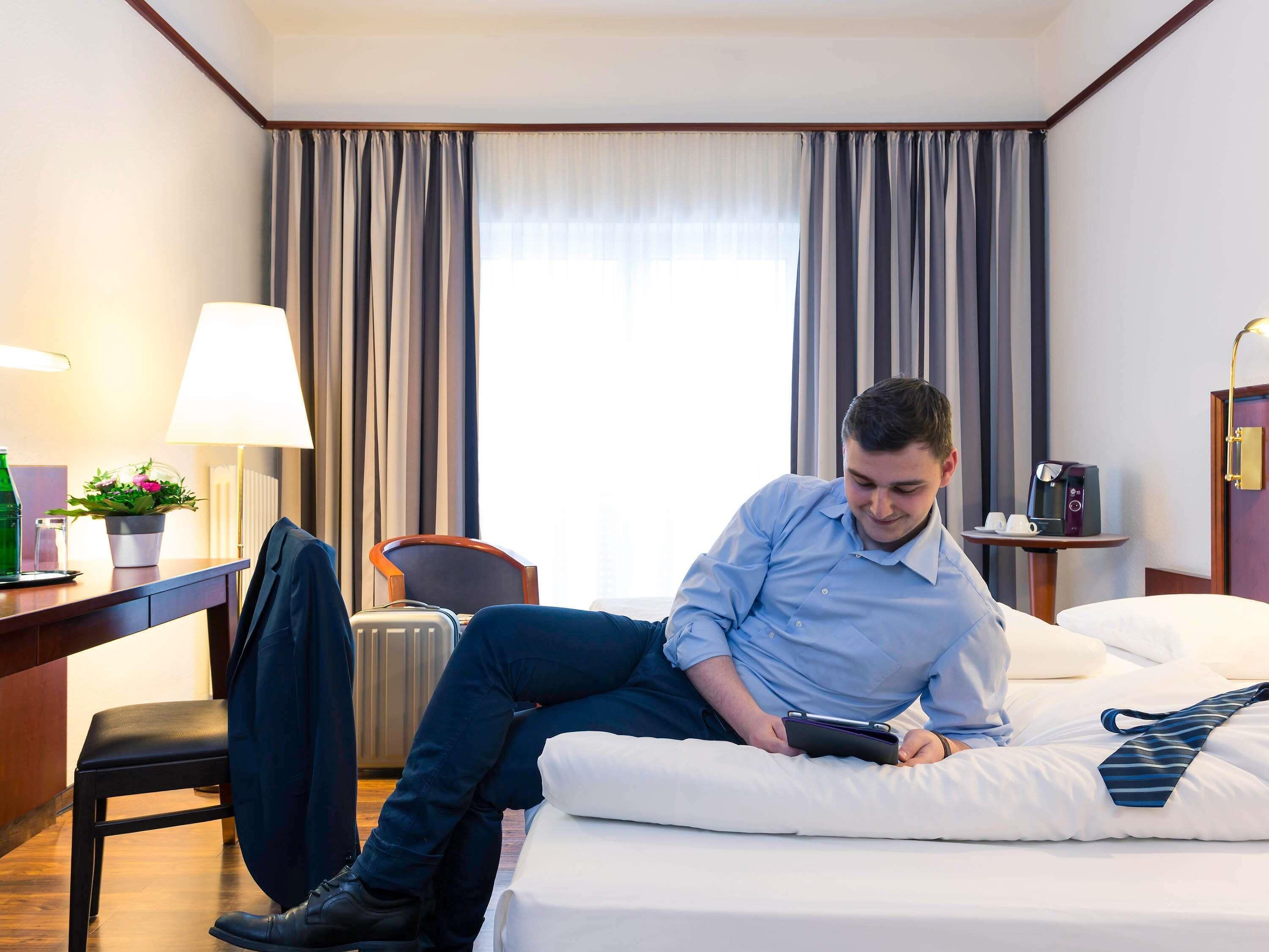 Et hotelværelse med seng, skrivebord, stol, lampe, kaffemaskine og et vindue med gardiner.