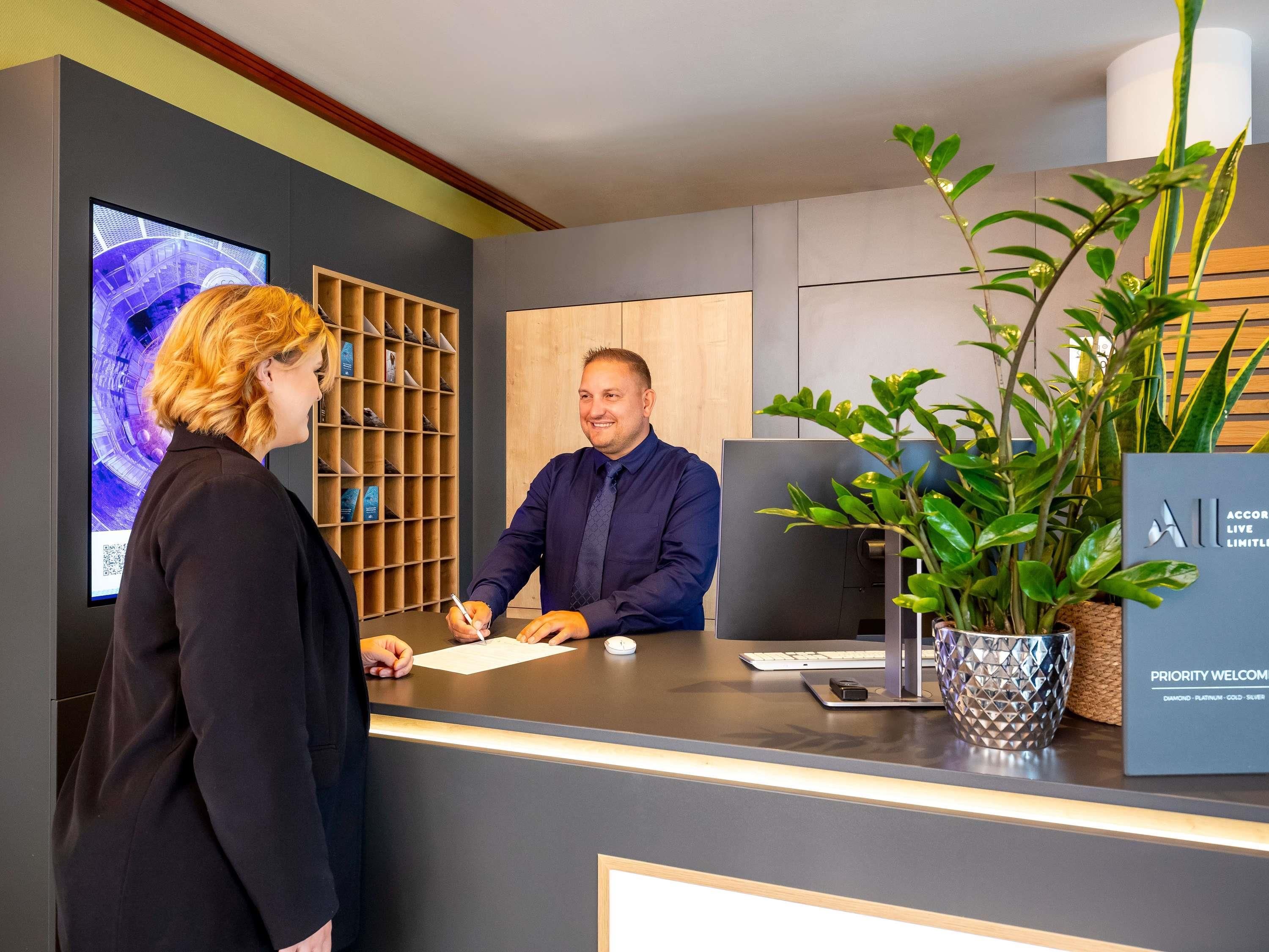 A reception desk with a digital screen, a plant, and a sign indicating priority welcome.