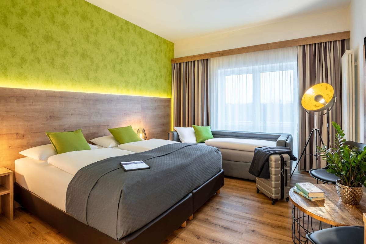 A hotel room with a bed, bedside table, lamp, and a view of the outdoors.