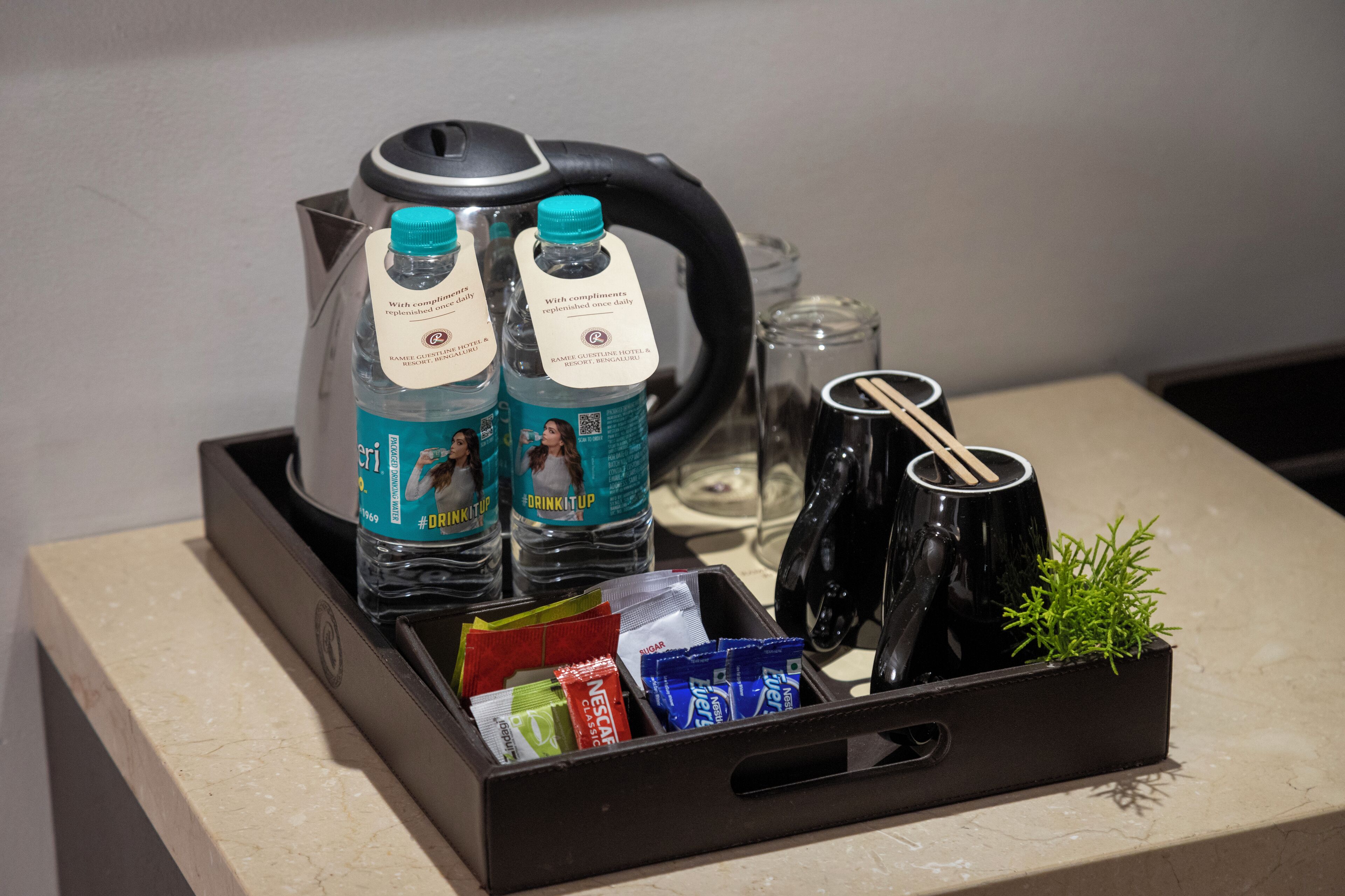 A tray with a kettle, two water bottles, a glass, a small plant, and various packaged items.