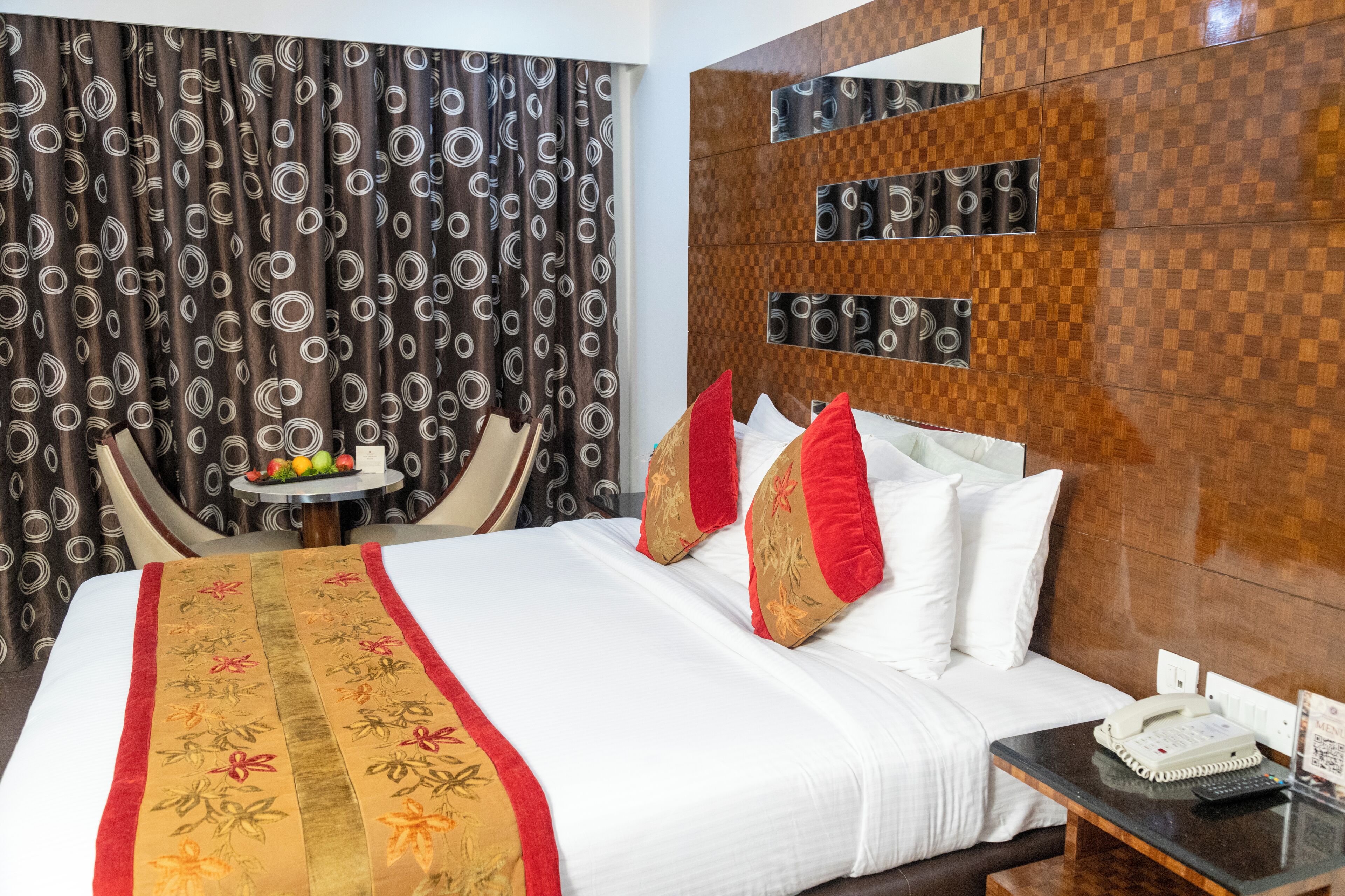 Habitación de hotel con una cama grande, una mesita de noche con un teléfono y un control remoto, una pequeña mesa redonda con fruta y un florero, y una pared con diseño a cuadros y motivos circulares.