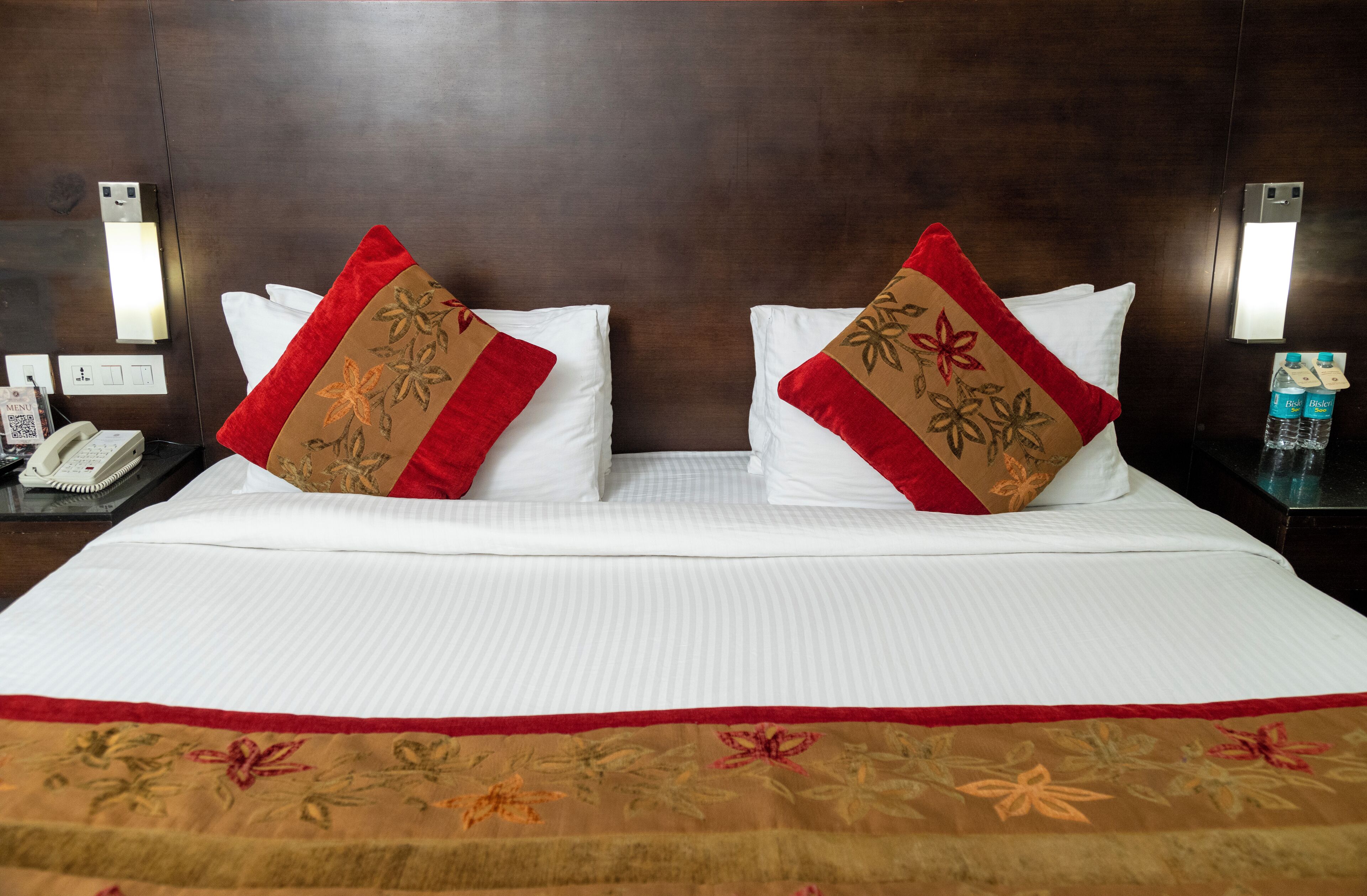 Una habitación de hotel con una cama grande, dos almohadones decorativos, una mesita de noche con un teléfono y un buró con agua embotellada.