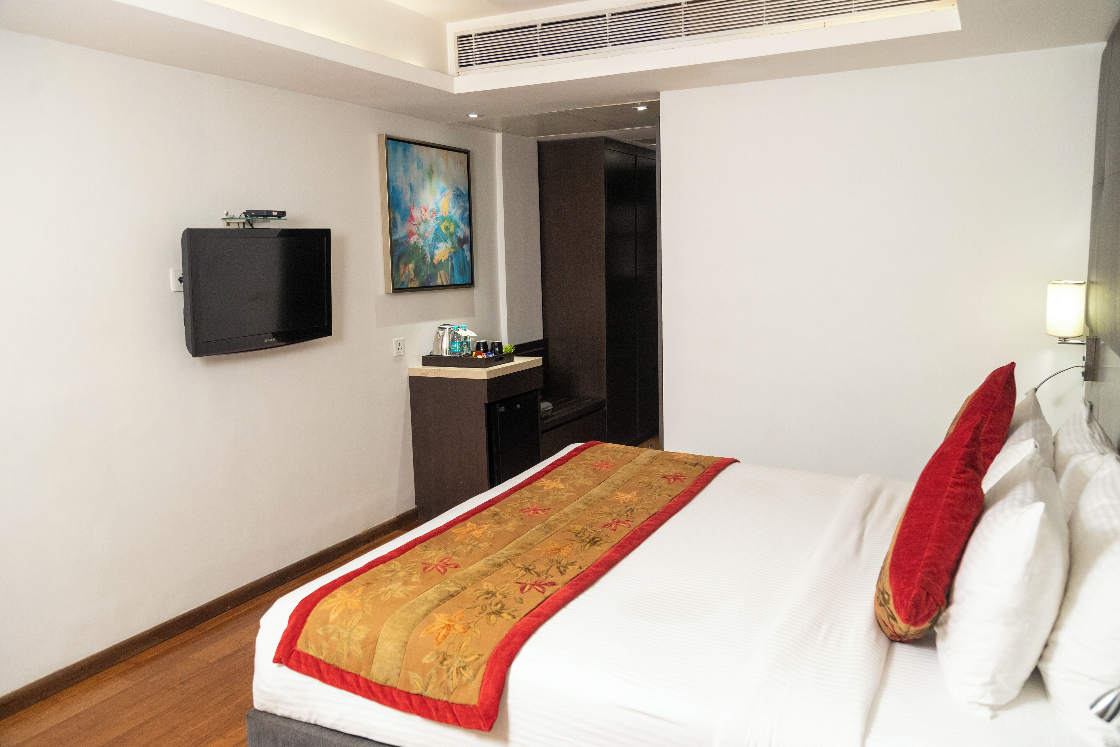 A hotel room with a bed, a flat-screen TV mounted on the wall, a painting, a desk with a lamp, and a door leading to another room.